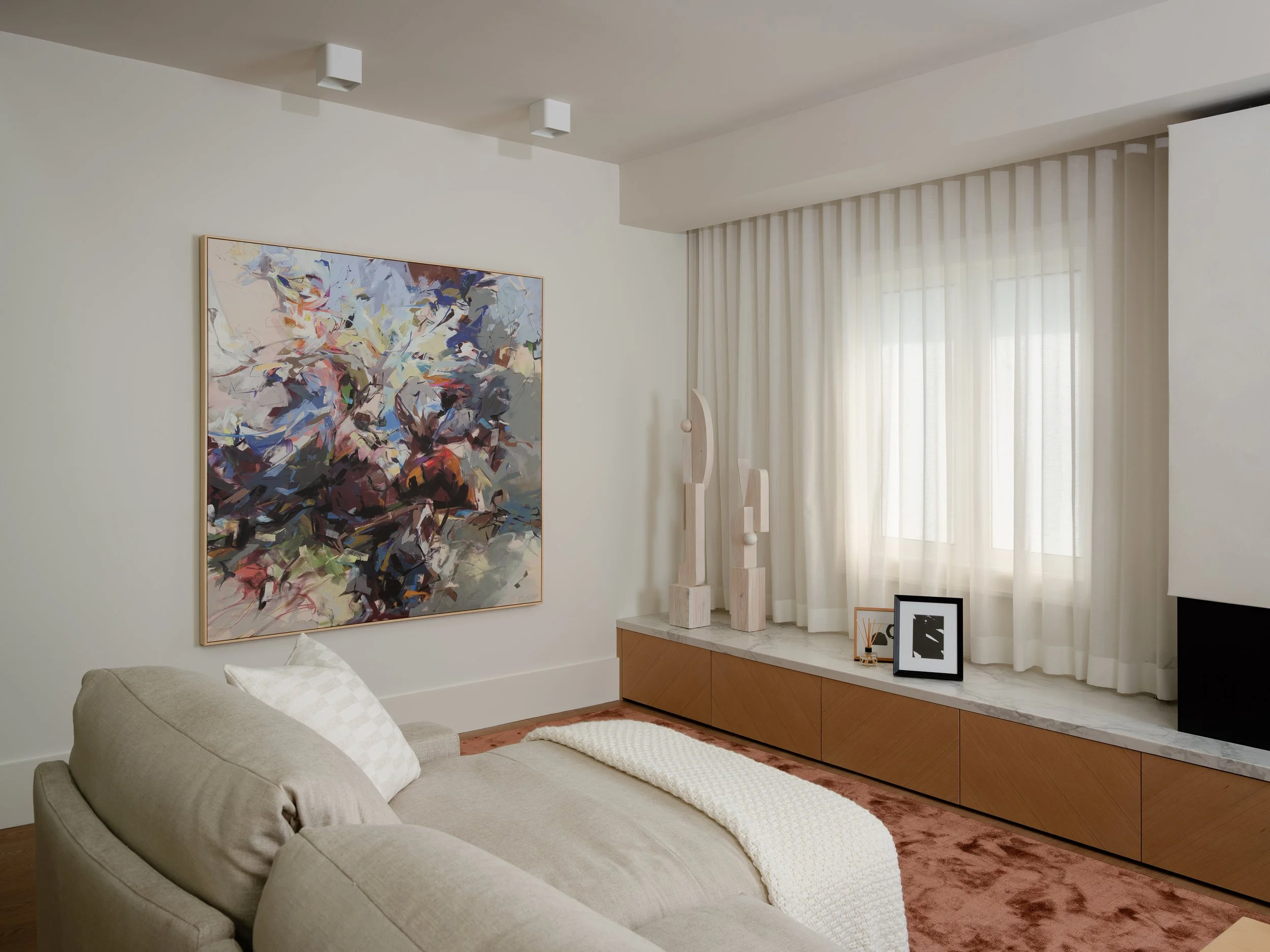 Living room corner with beige sofa, abstract wall art, white curtains, wooden cabinet with decor, and framed photos.