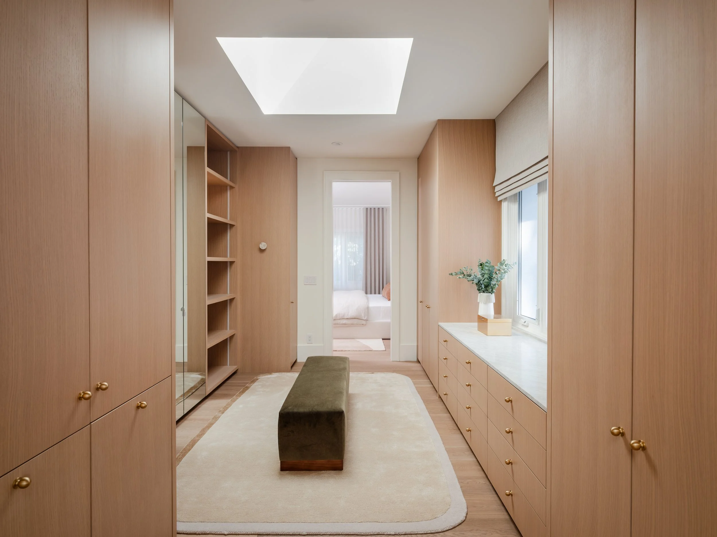 Interior view of a room with wooden cabinets on both sides, a window with a outside view, a beige area rug, a bench in the middle, a tall mirror, and a doorway leading to a bedroom with a bed, window with curtains, and a nightstand.