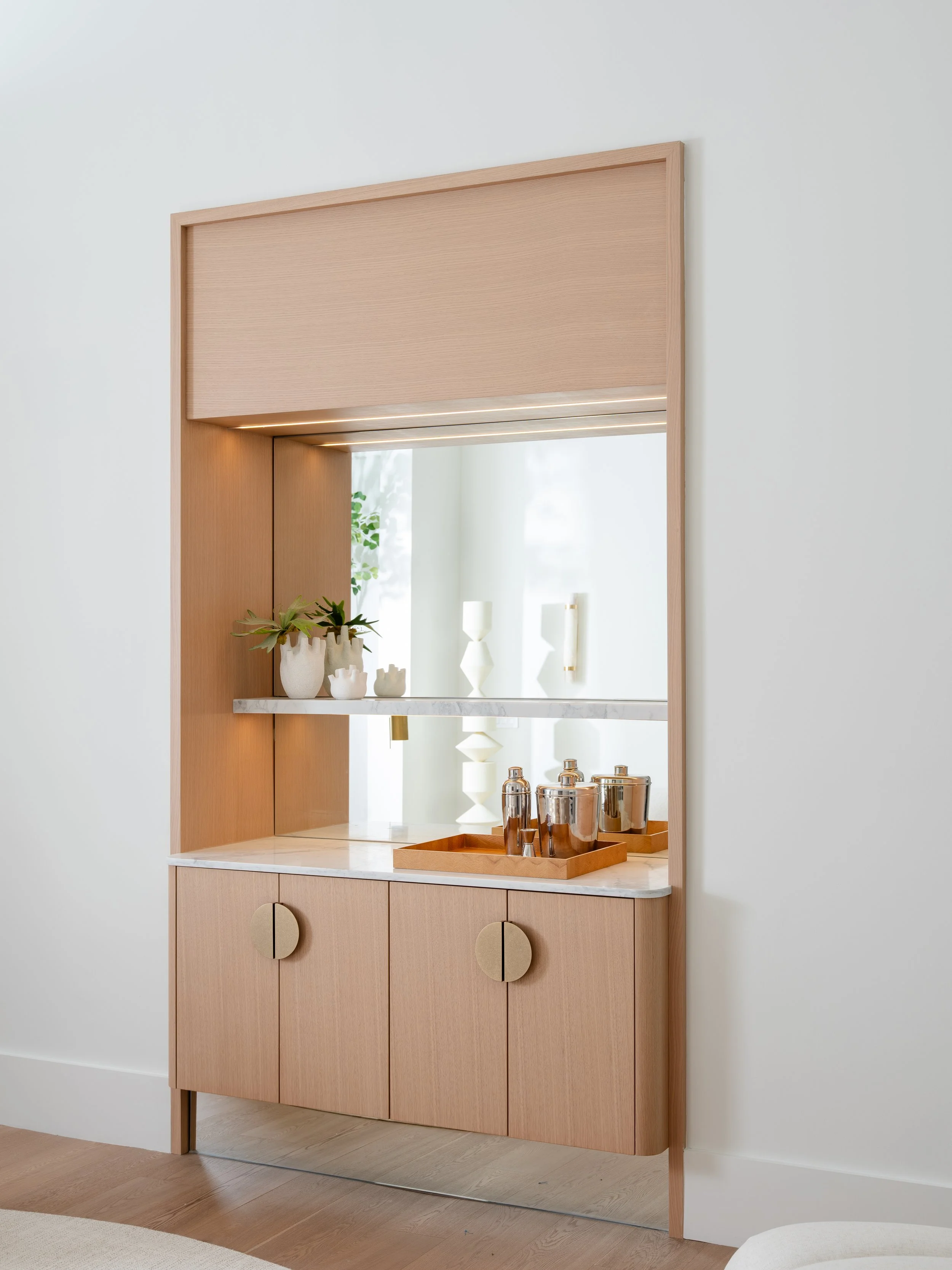A light wood cabinet with a marble top, decorated with white vases and metallic cocktail shakers, against a white wall with a mirror reflecting a lamp and a plant.