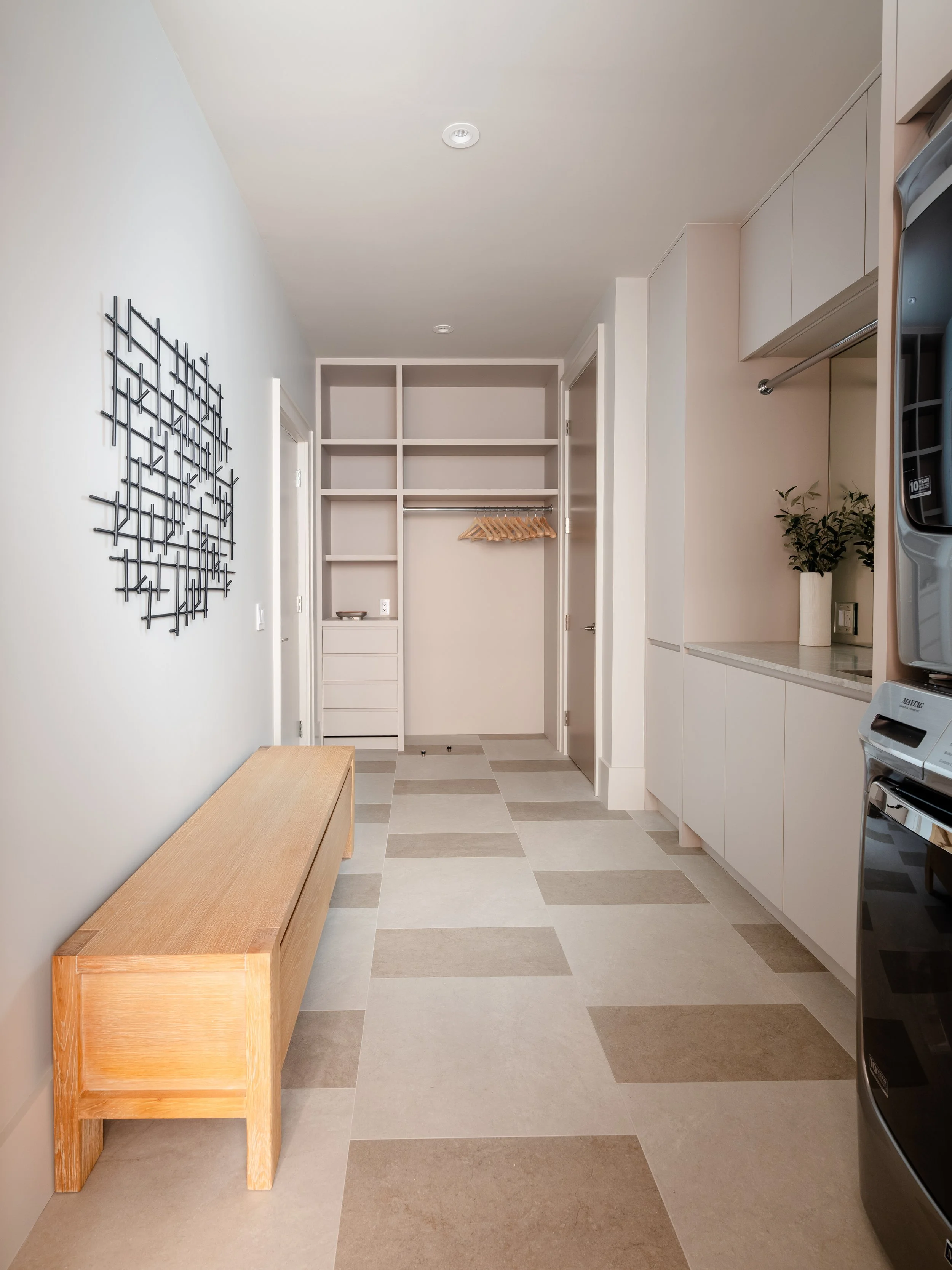 A laundry room with white cabinets, a beige wall, a wooden bench, and a patterned tile floor. The room has a decorative black metal wall hanging, a potted plant, and a laundry area with washer and dryer units.