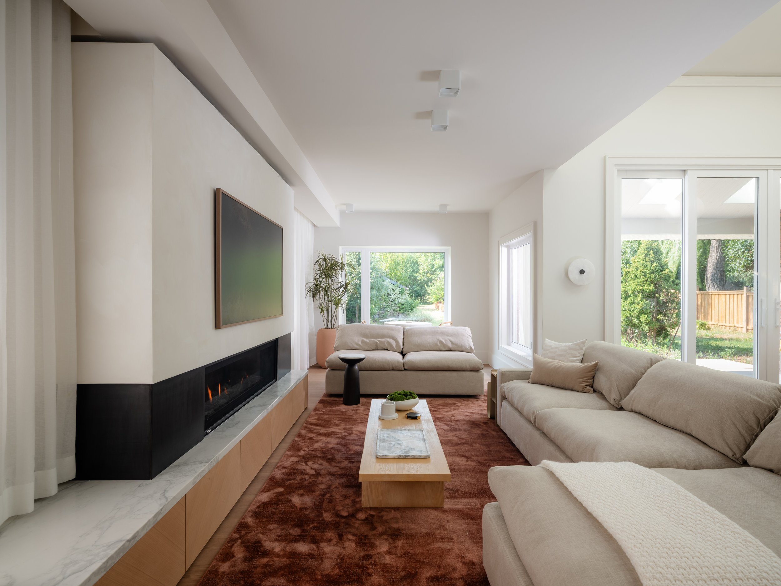 A modern living room with a beige sofa, a wooden coffee table, and a large flat-screen TV on a black and beige media unit. The room has large windows with views of green trees outside and a red rug on the floor.