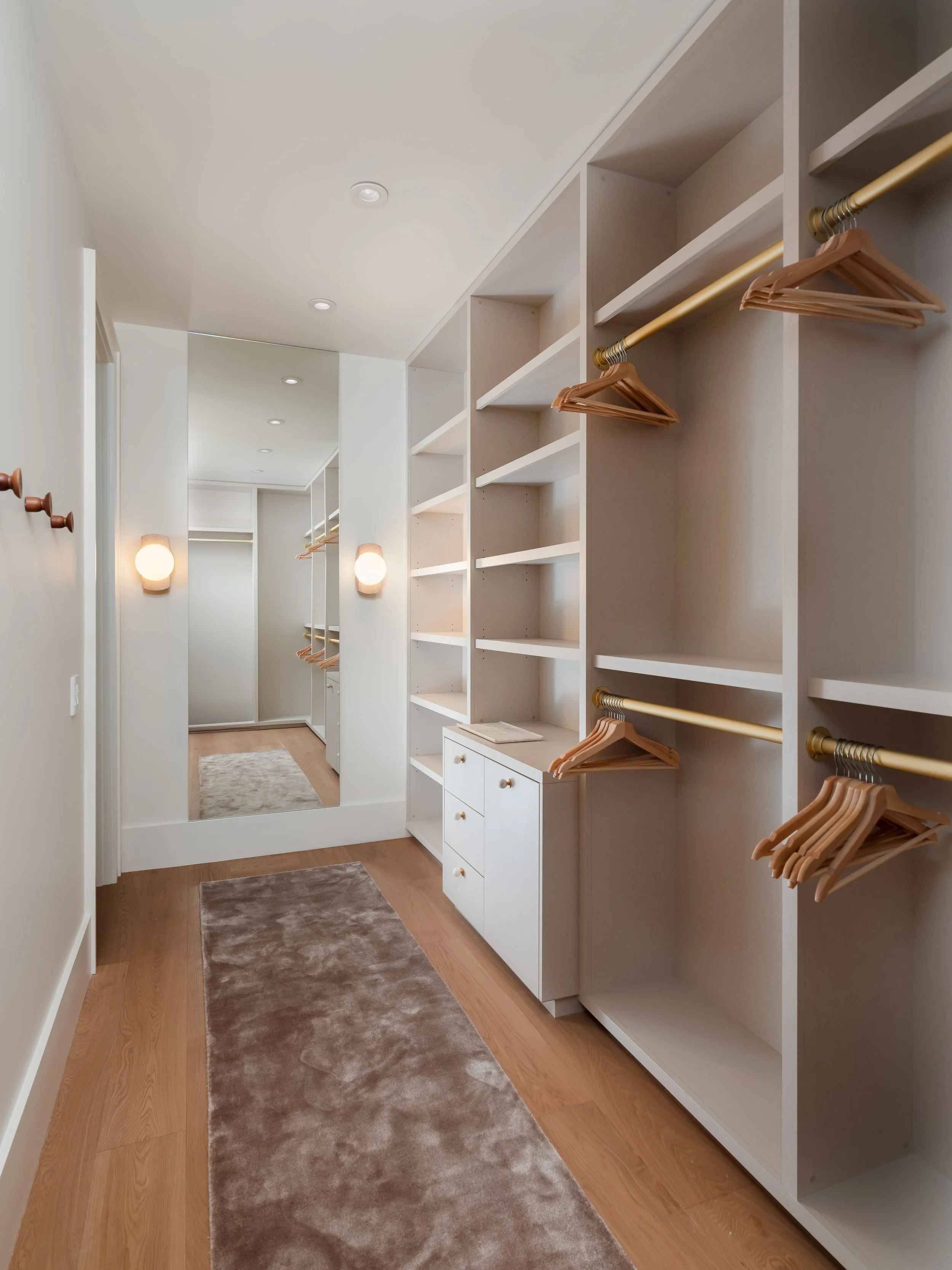Empty walk-in closet with white built-in shelves and hanging rods, beige wall-mounted lamps, light wood flooring, and a mirror at the end of the space.