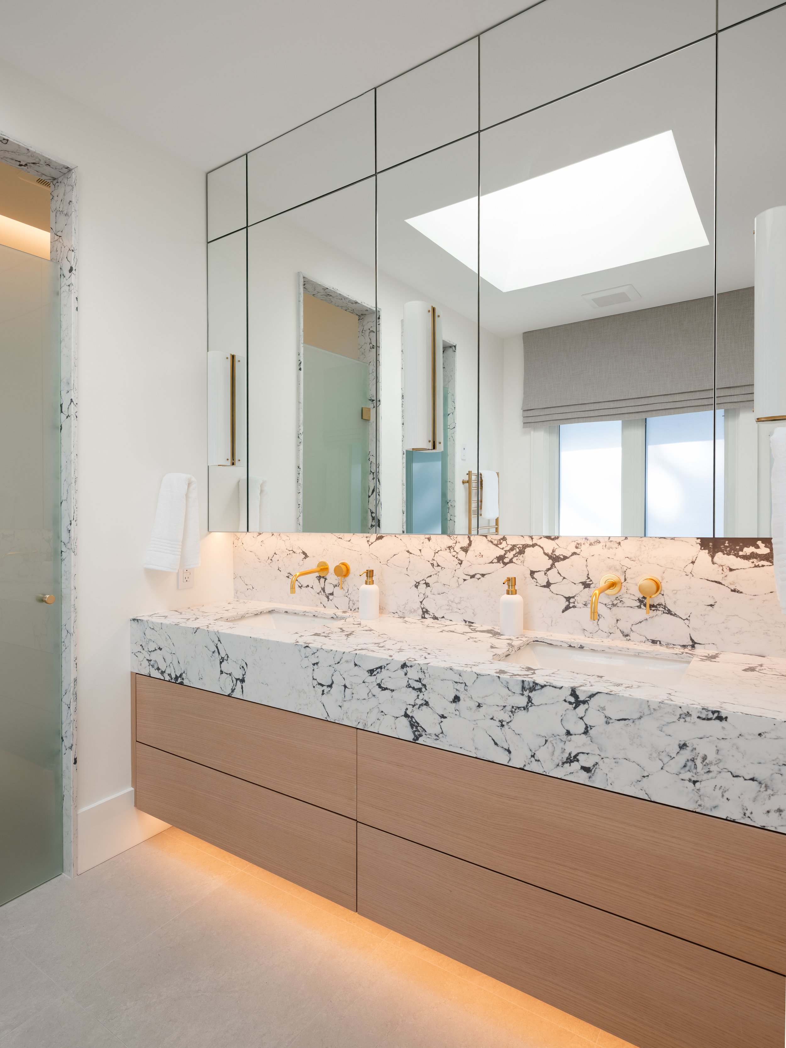Modern bathroom with a marble vanity, gold fixtures, large mirror, and a window with a beige roman shade.