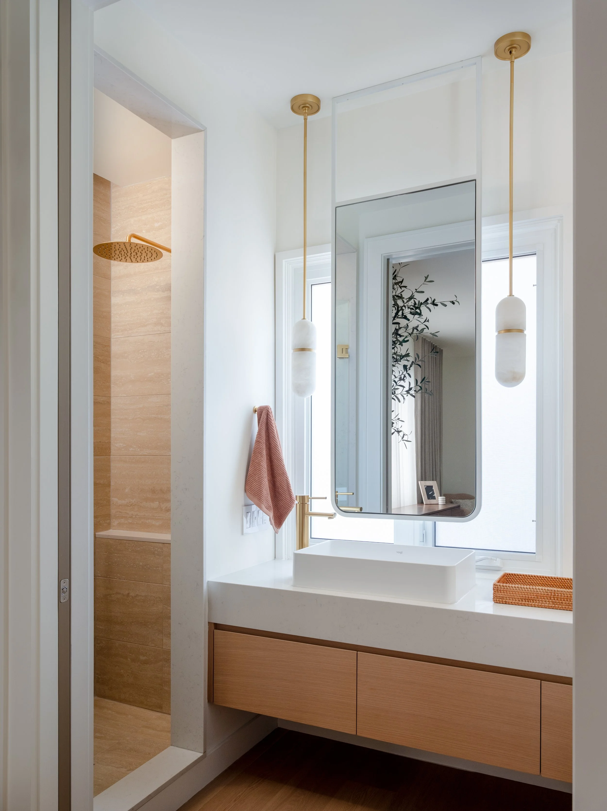 Modern bathroom with a floating wooden vanity, a rectangular vessel sink, large mirror, two hanging pendant lights, and a window. The shower area with a rain shower head is visible to the left.