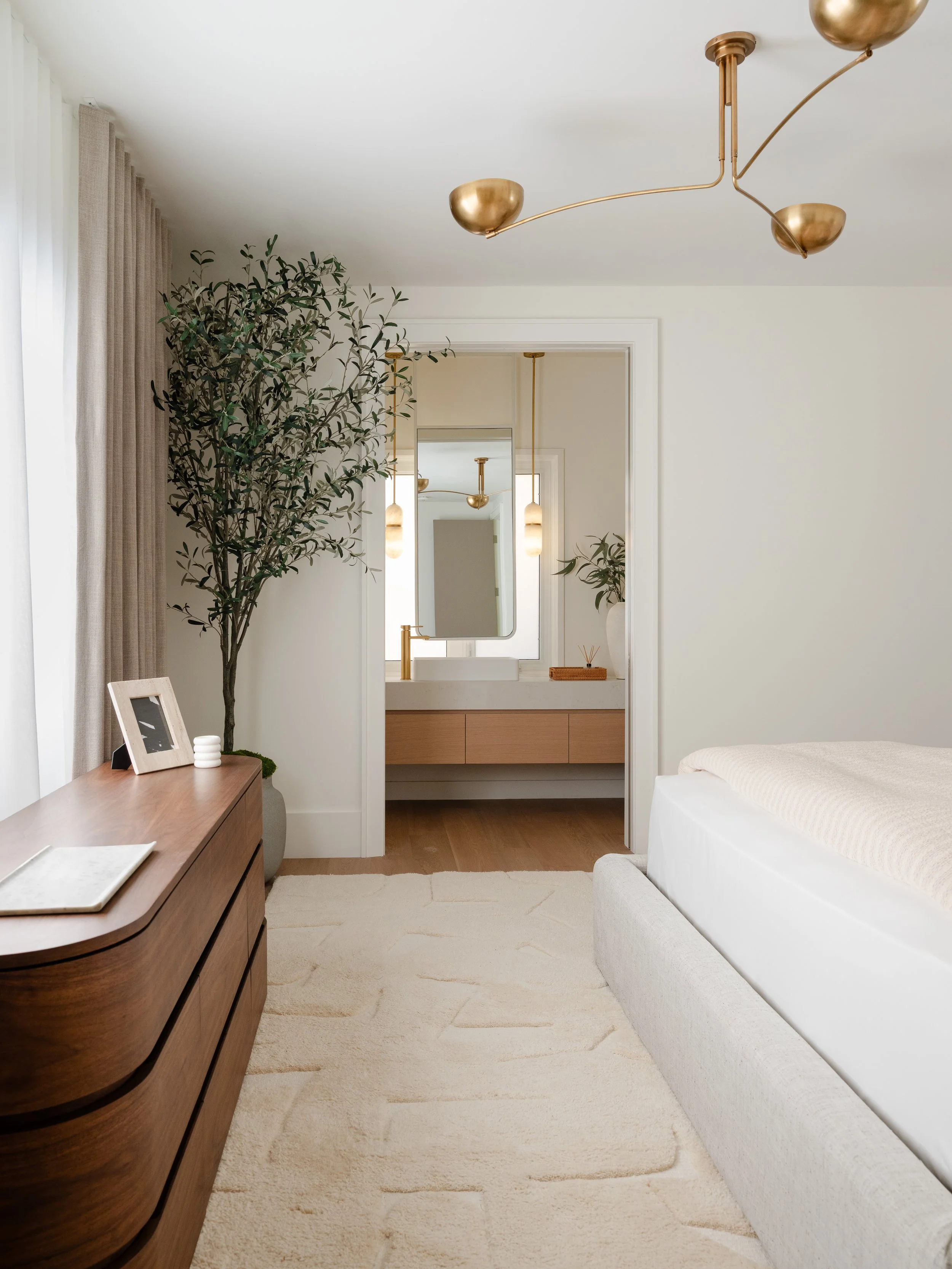 A bedroom with a white bed, a wooden dresser with a photo frame and a small white jar, a beige rug, a large potted plant, beige curtains, and modern gold ceiling light fixtures.