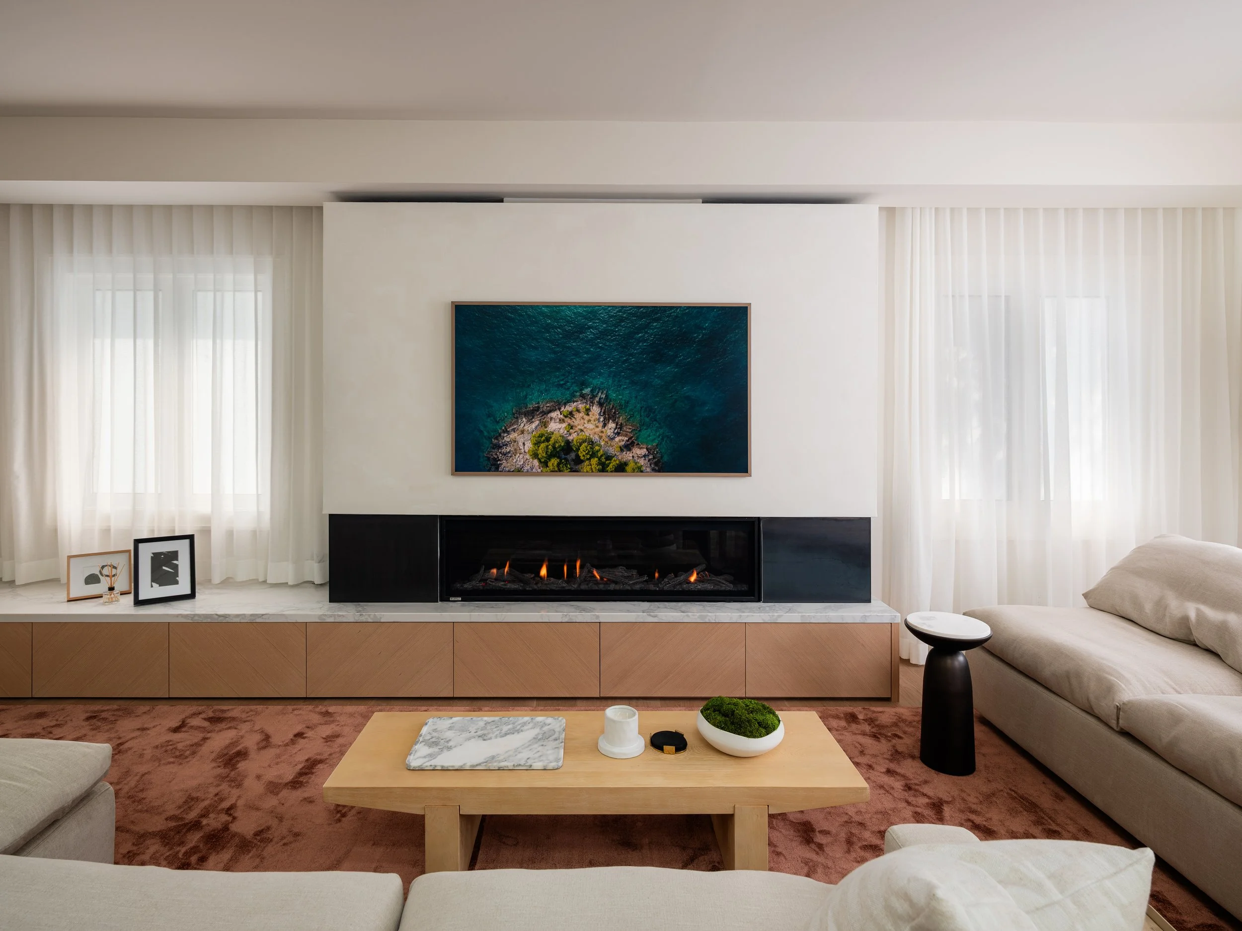 Living room with a modern fireplace, framed artwork, and large windows with white curtains.