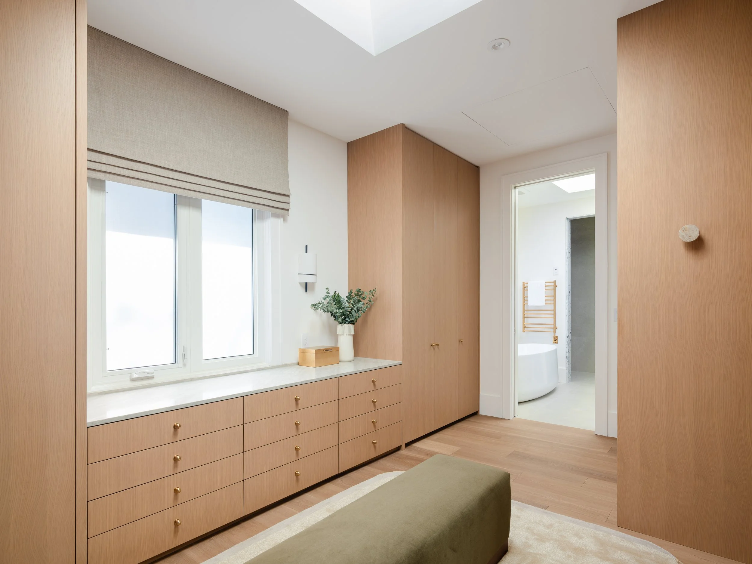 A room with light wood cabinetry, a large window with a beige Roman shade, a white bathtub visible through a doorway, a small plant, and a beige rug and ottoman.