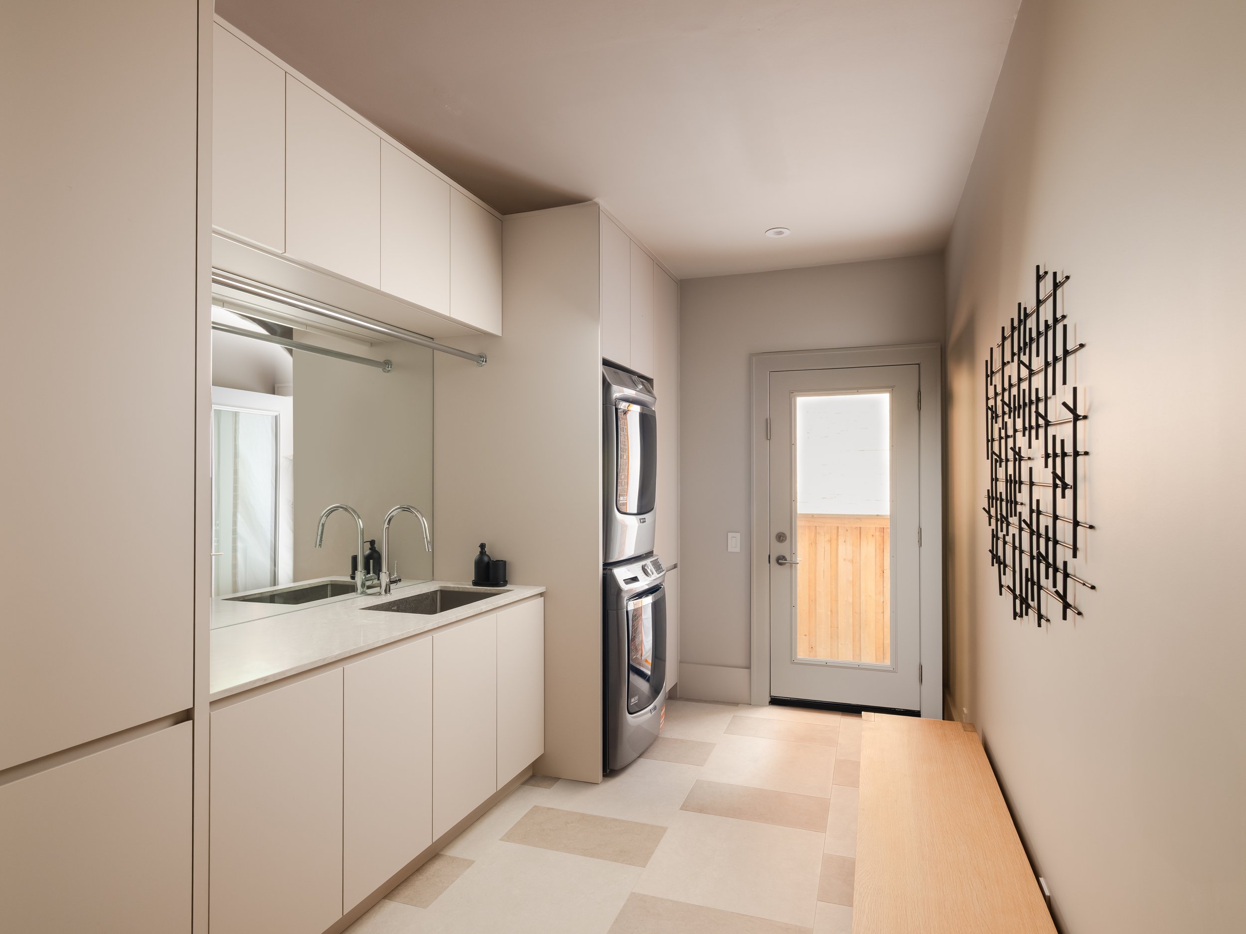 Modern laundry room with white cabinets, a stainless steel washer and dryer, a double sink with a faucet, and a window with a door leading outside.
