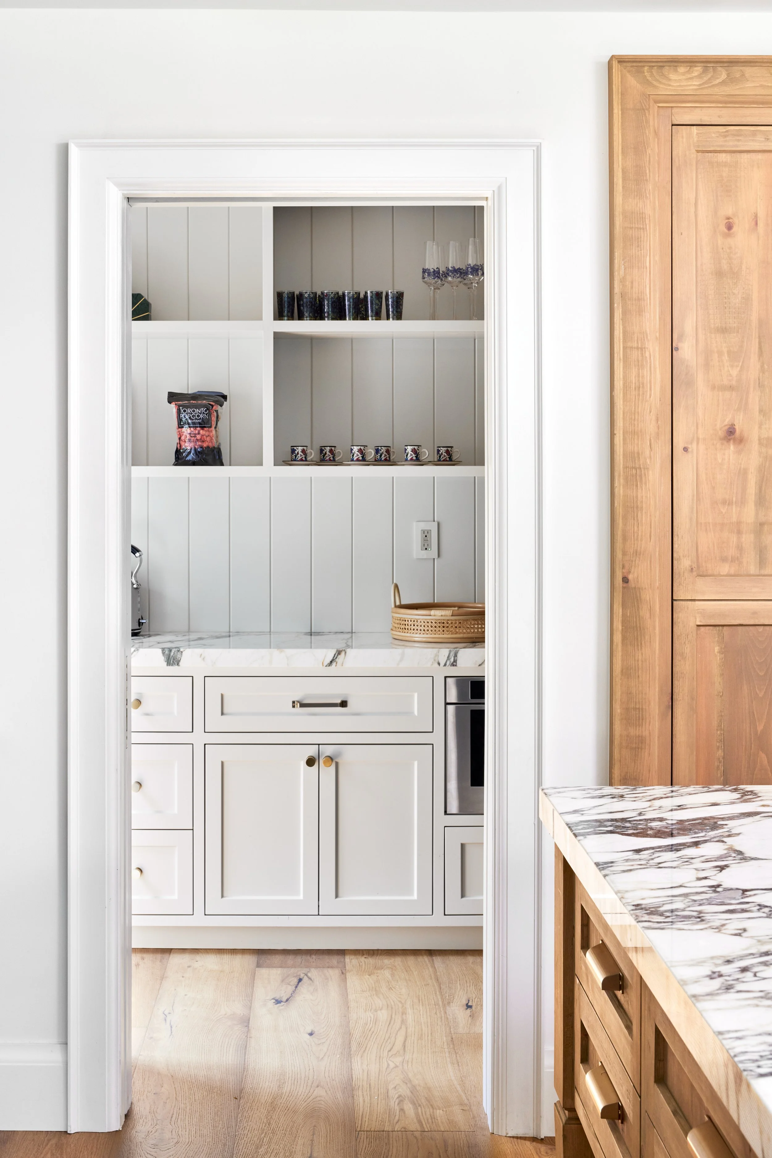 View of a white kitchen nook with open shelving, containing black and printed cups, clear champagne flutes, a bag of Toronto popcorn, and a small tray. The space features a white paneled wall, a marble countertop, and light wood cabinetry to the righ