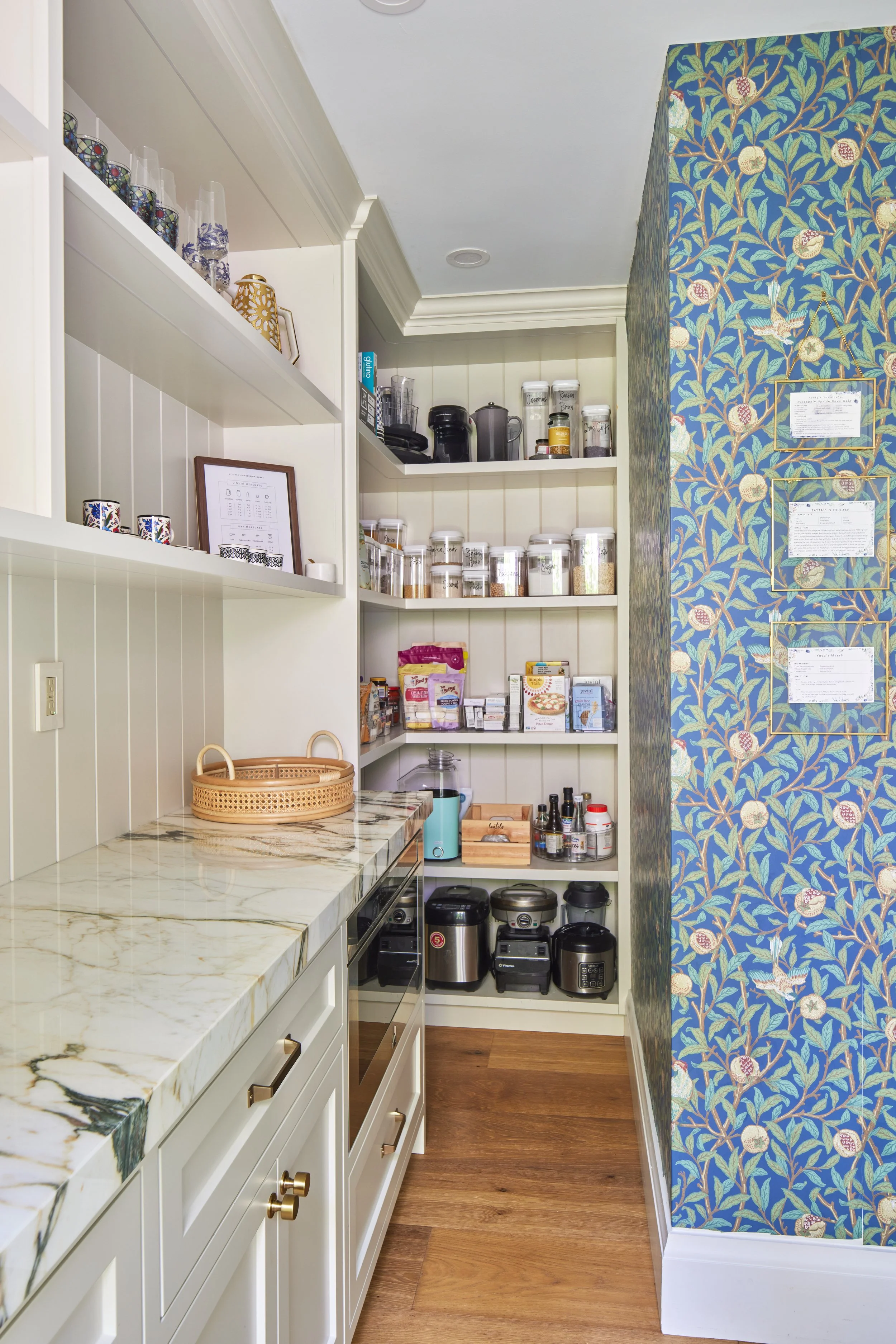 A view of a kitchen corner with white cabinetry, a marble countertop, open shelves containing jars and kitchen appliances, and a blue wallpapered wall with a pomegranate and bird pattern.