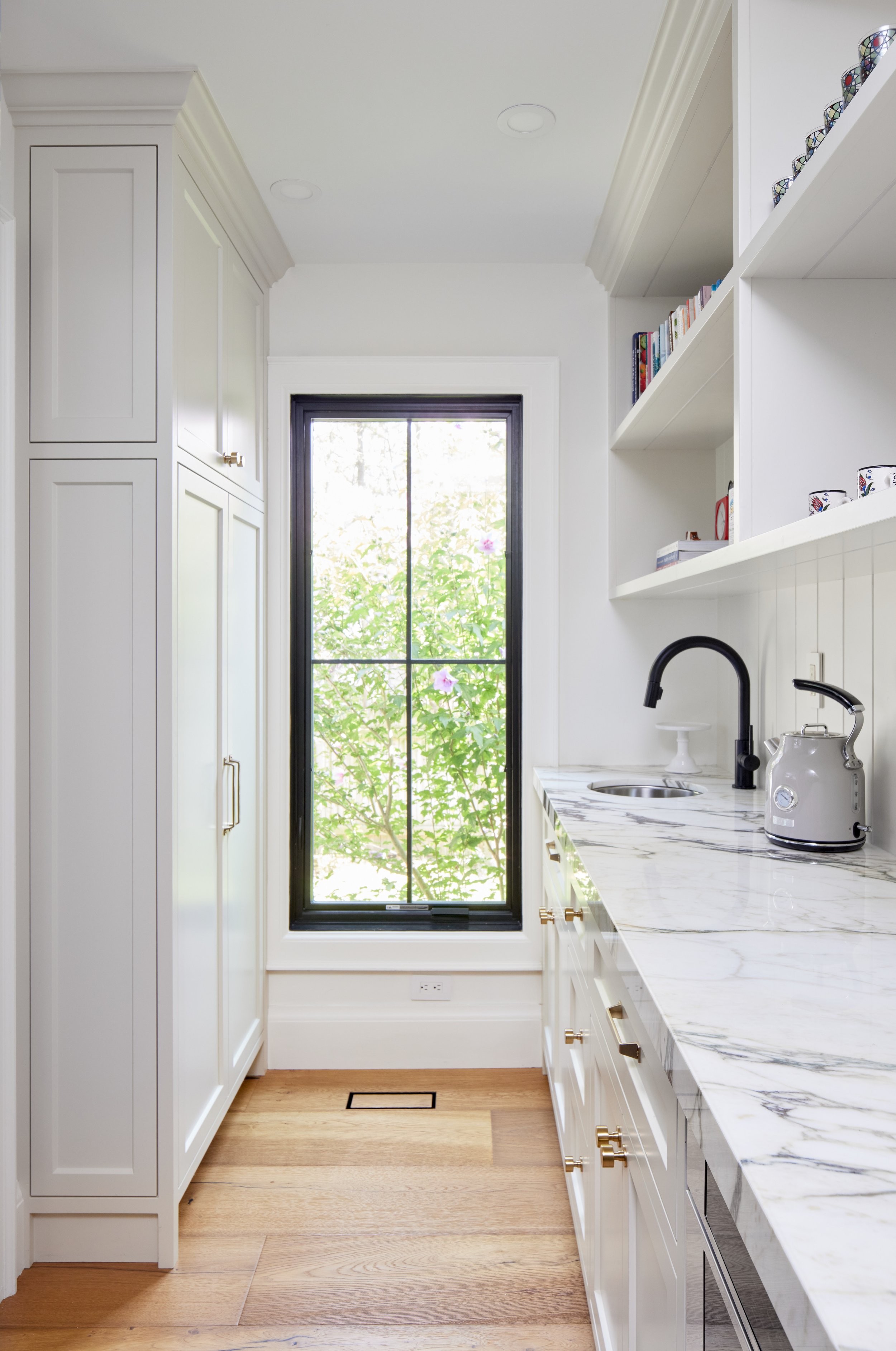 A modern kitchen with white cabinets, a marble countertop, a black faucet, a window with greenery outside, and light wooden flooring.