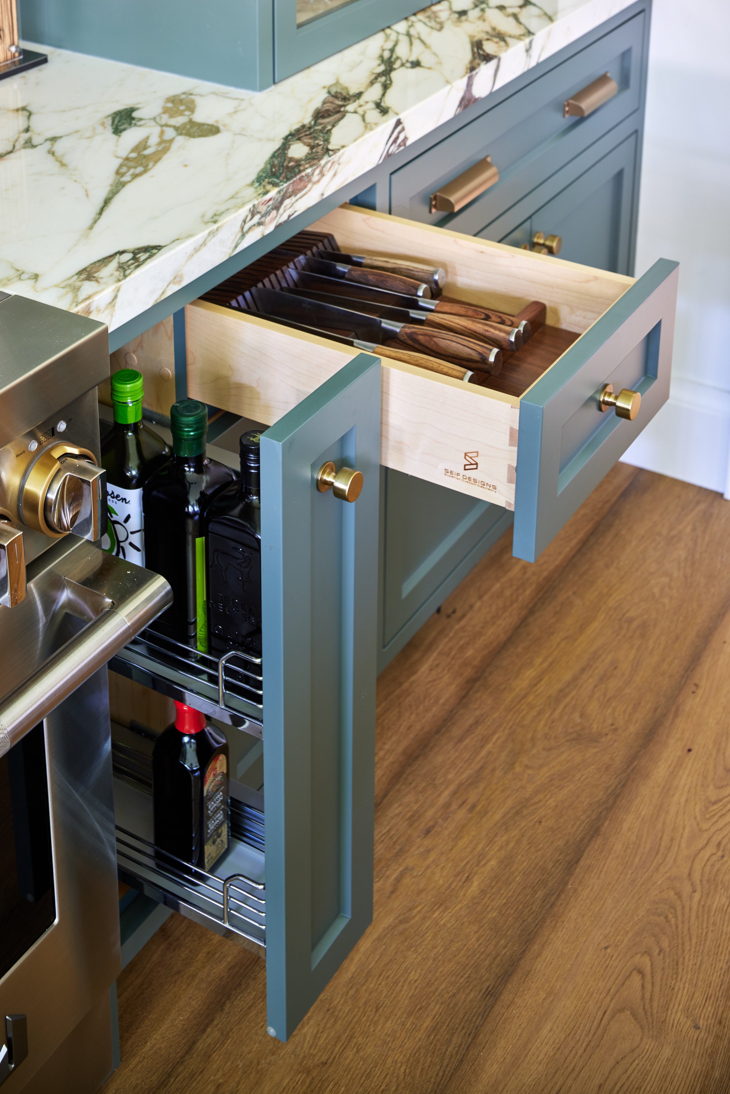 High-end custom modern kitchen in Mississauga, Oakville, and Toronto, Ontario. Features trends color for 2024, luxurious gold hardware, a marble countertop, an elegant kitchen sink, custom shelving with a pocket door, and efficient kitchen and drawer
