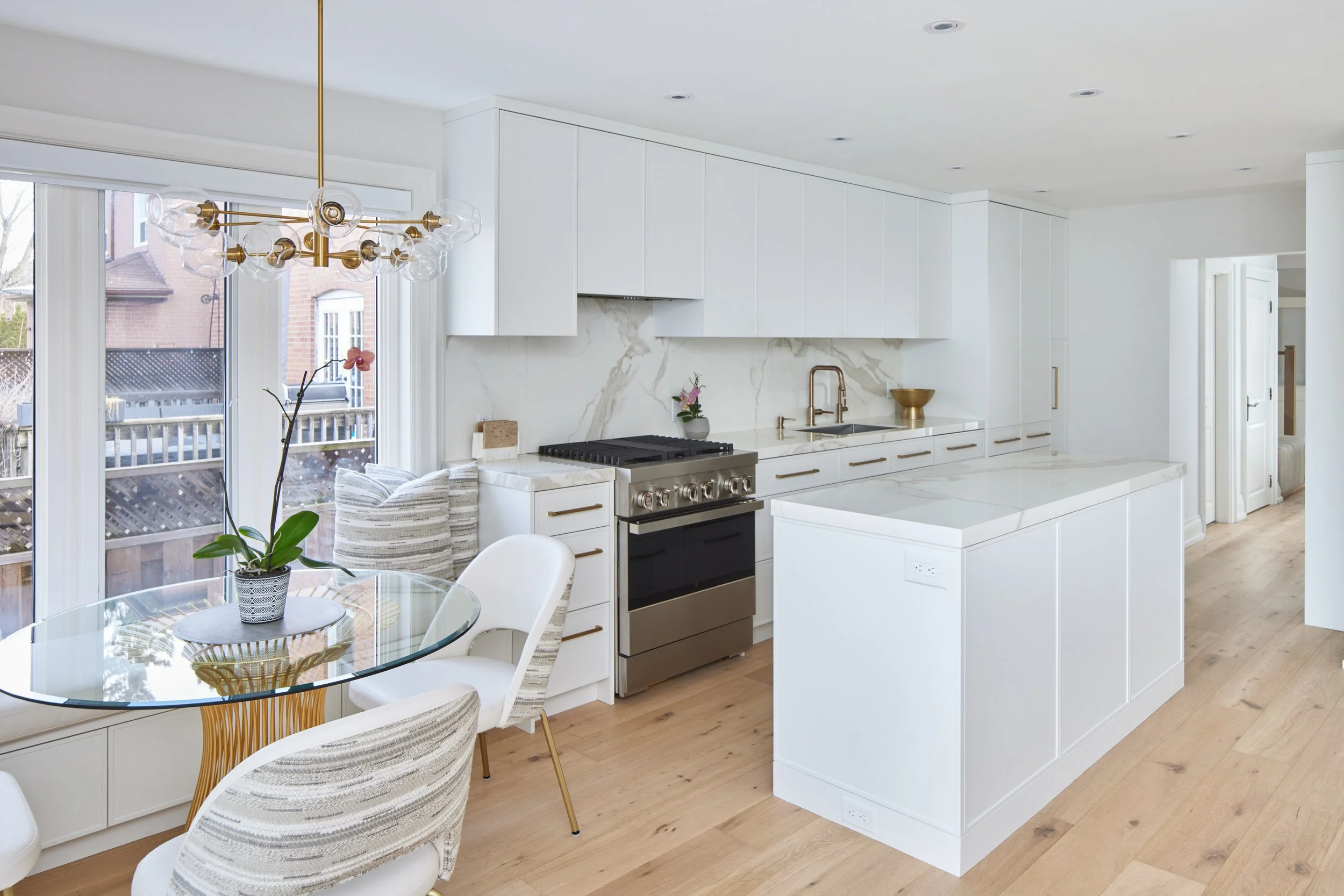 Explore This Beautiful Kitchen: See a modern kitchen with clean white cabinets and beautiful marble. It's well-designed with gold handles and light wood floors that make the space look bright and welcoming. Nearby, a window bench in the dining area o