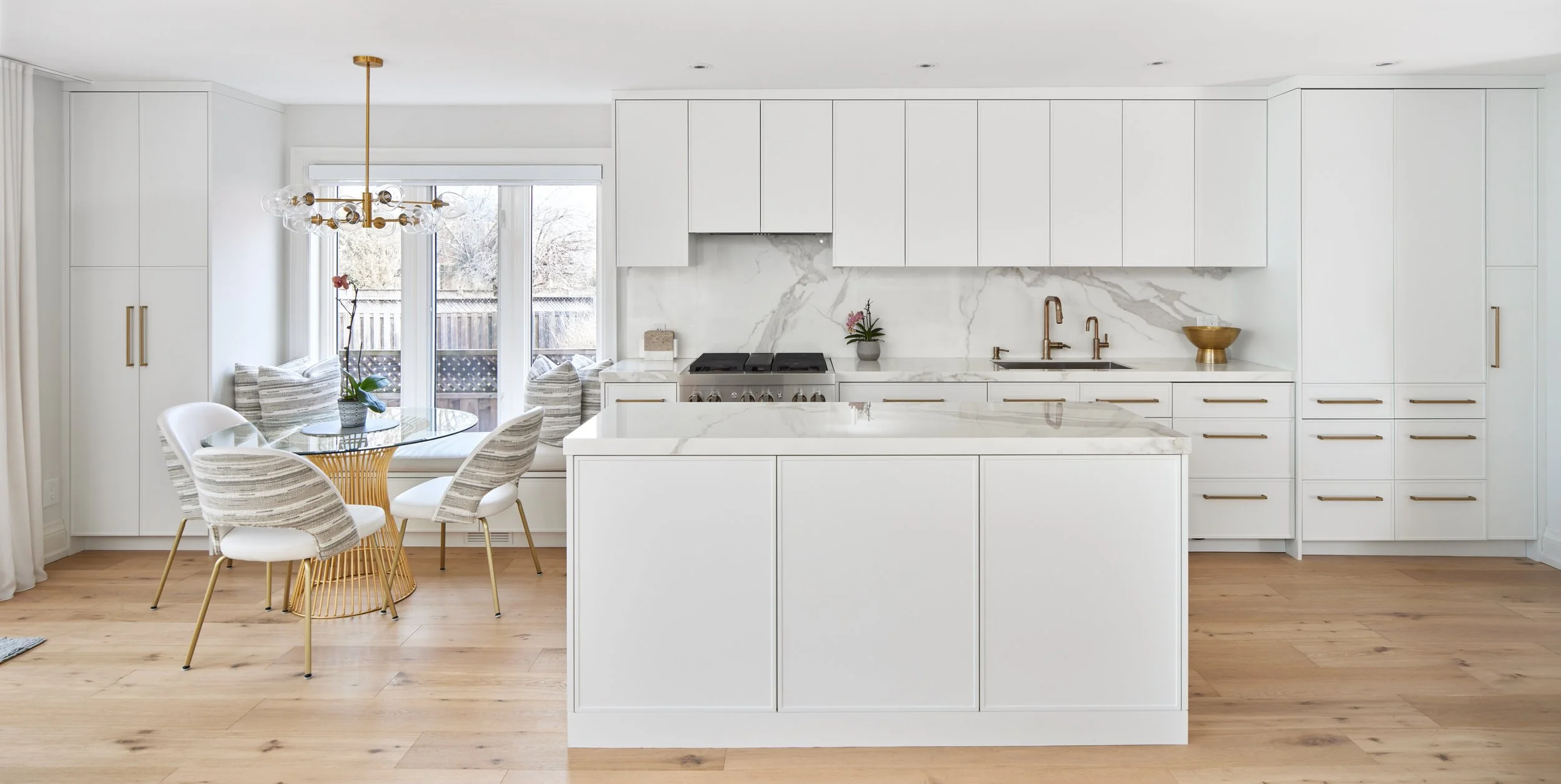 Explore This Beautiful Kitchen: See a modern kitchen with clean white cabinets and beautiful marble. It's well-designed with gold handles and light wood floors that make the space look bright and welcoming. Nearby, a window bench in the dining area o