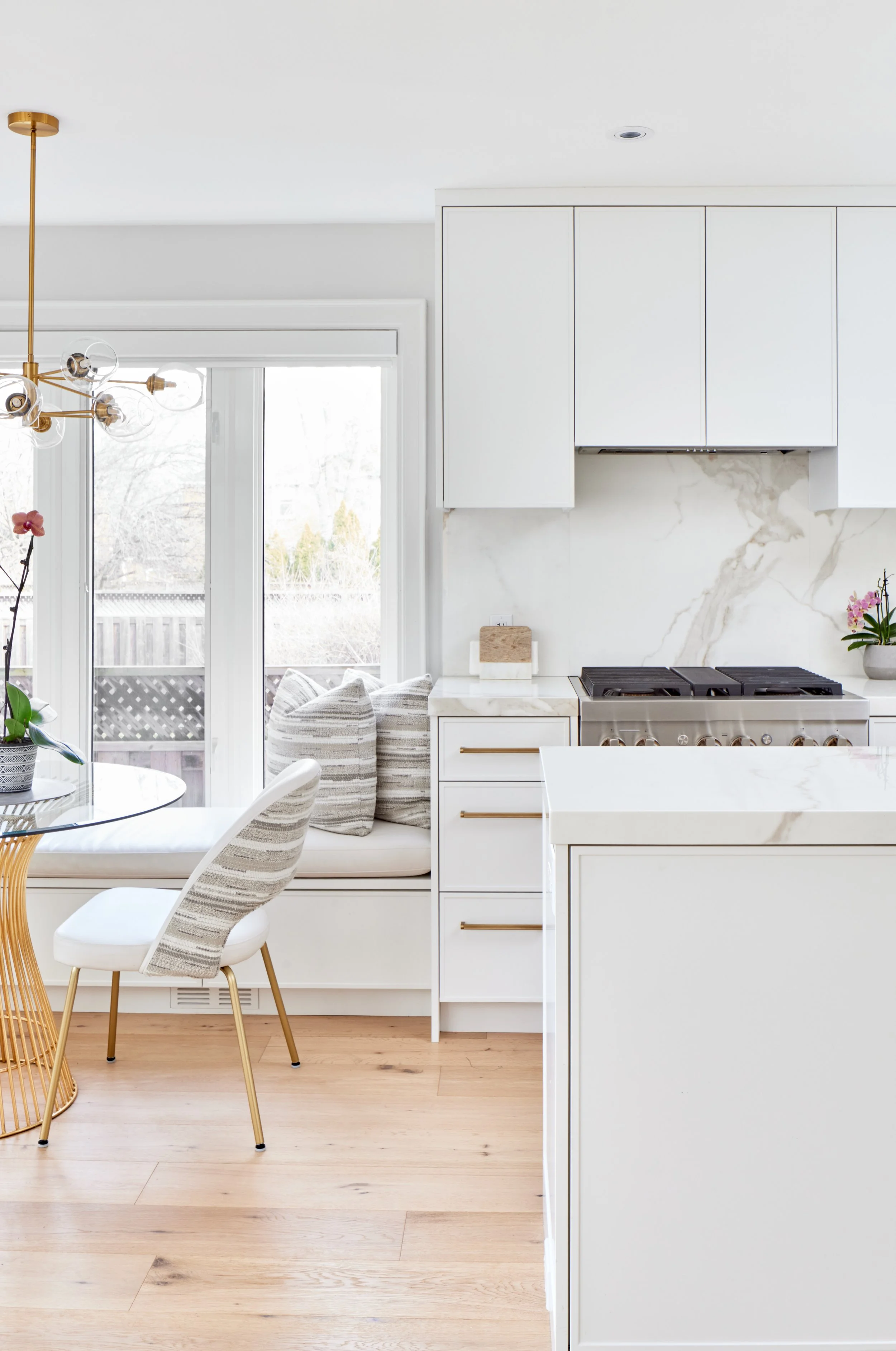 Explore This Beautiful Kitchen: See a modern kitchen with clean white cabinets and beautiful marble. It's well-designed with gold handles and light wood floors that make the space look bright and welcoming. Nearby, a window bench in the dining area o