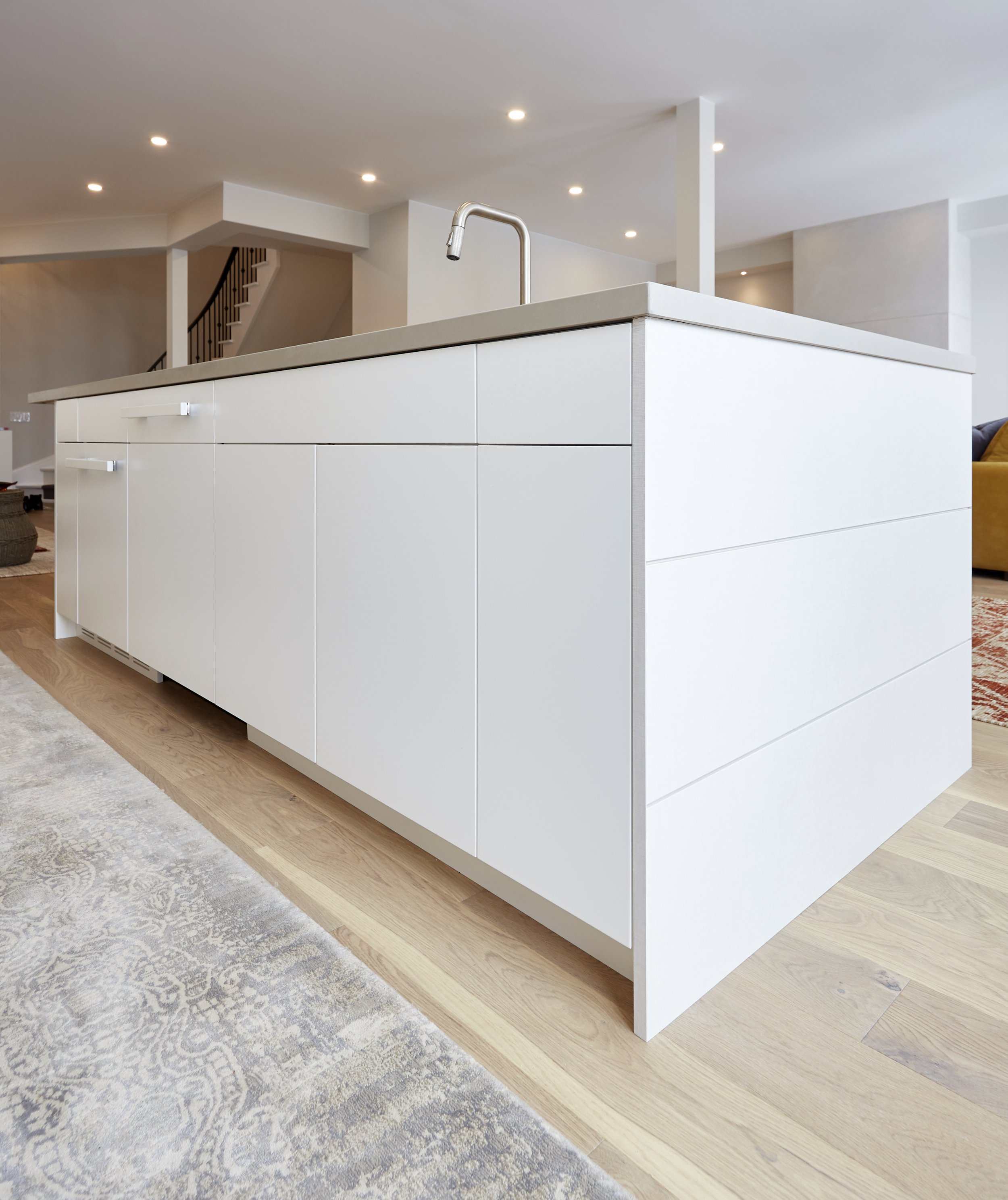 A modern kitchen with white cabinets and a large island,  the light wood floors match the simple and clean look of the kitchen. It's a practical space for cooking and has a few plants and a piece of art to add some character.  Serving kitchen cabinet