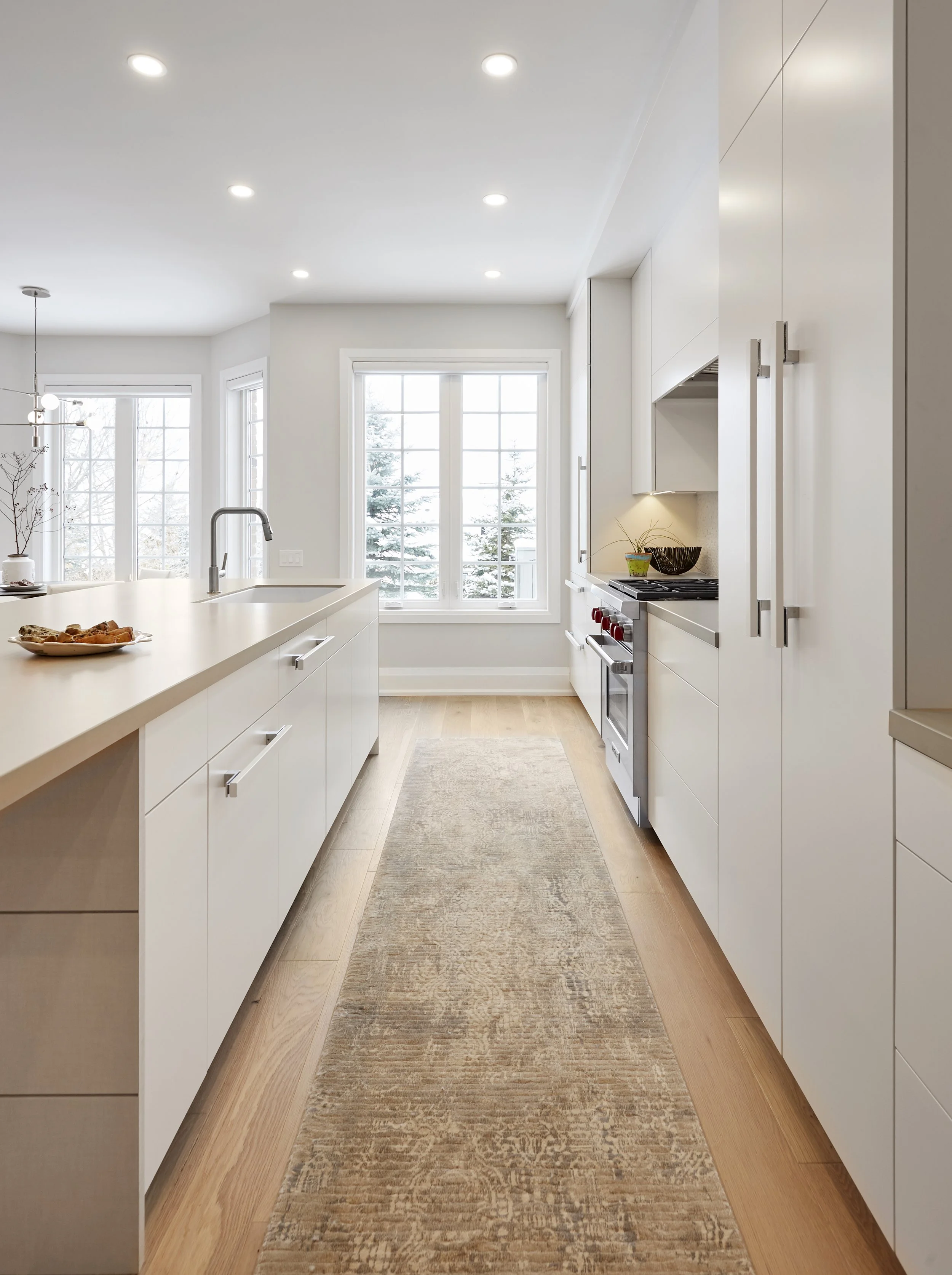 A modern kitchen with white cabinets and a large island,  the light wood floors match the simple and clean look of the kitchen. It's a practical space for cooking and has a few plants and a piece of art to add some character.  Serving kitchen cabinet