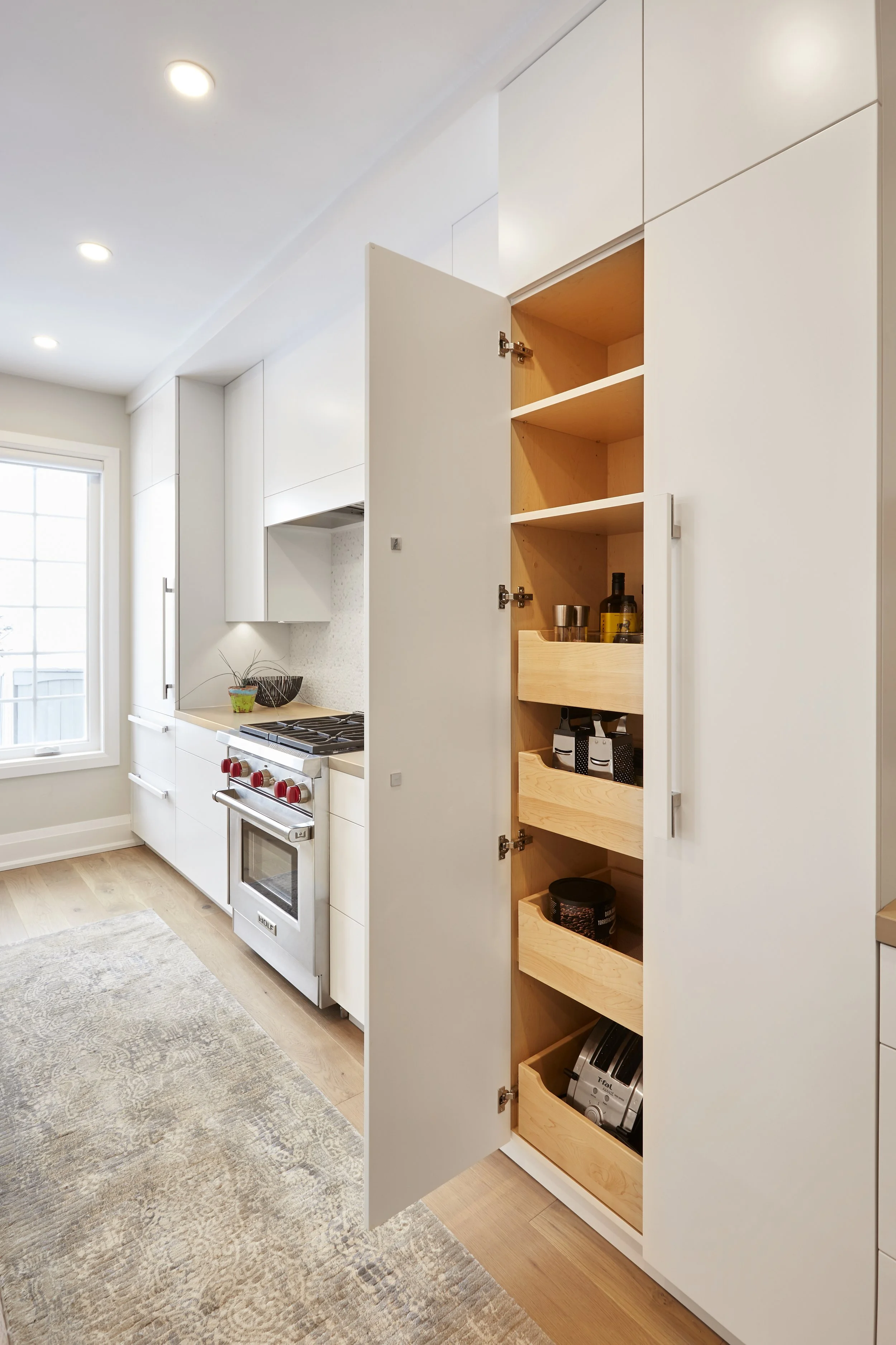 A modern kitchen with white with smart storage solutions. features a tall cabinet with open doors, revealing neatly organized shelves filled with cooking essentials, which makes finding and reaching items easy. A hidden compartment in the island that