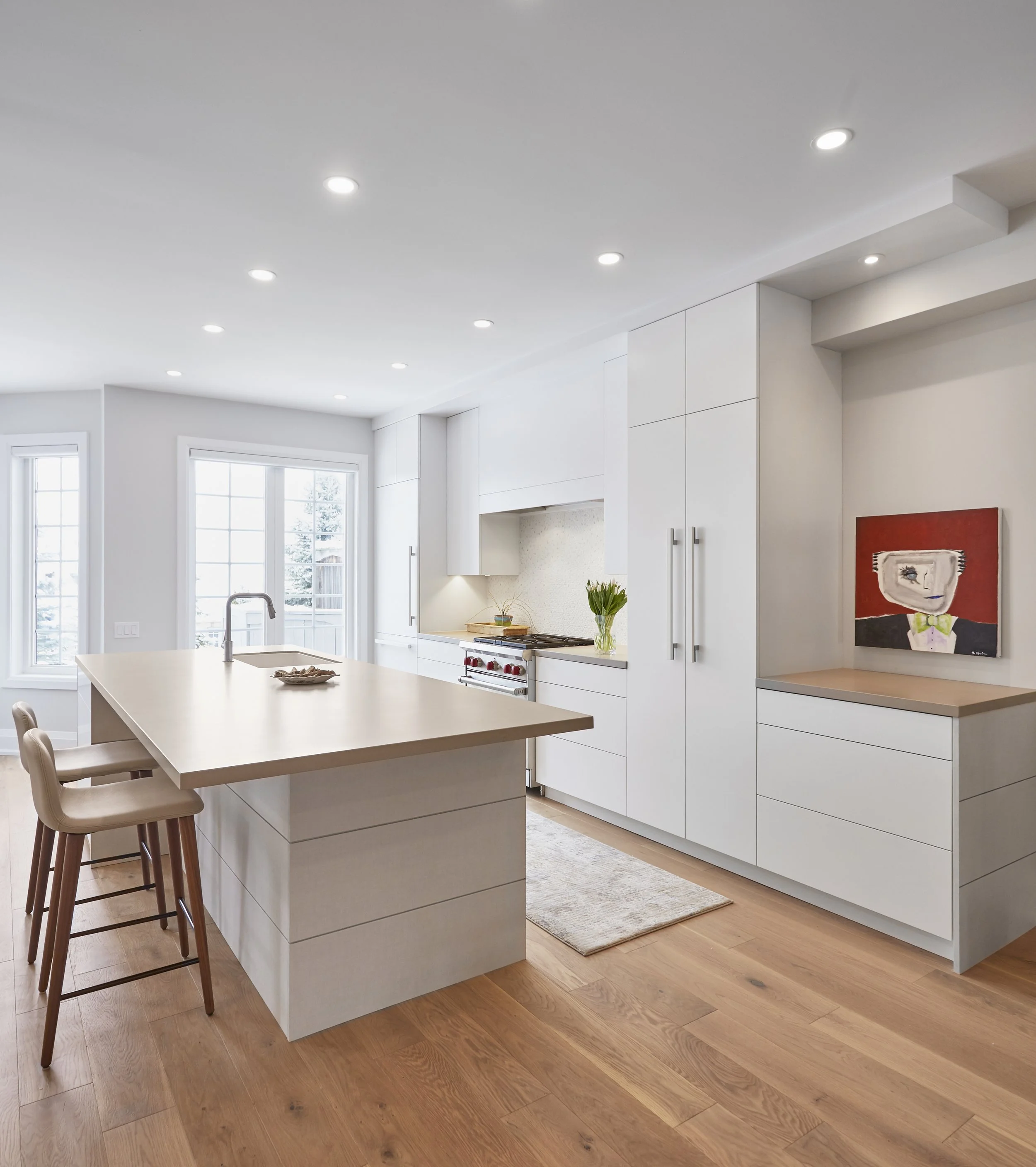 A modern kitchen with white cabinets and a large island,  the light wood floors match the simple and clean look of the kitchen. It's a practical space for cooking and has a few plants and a piece of art to add some character.  Serving kitchen cabinet