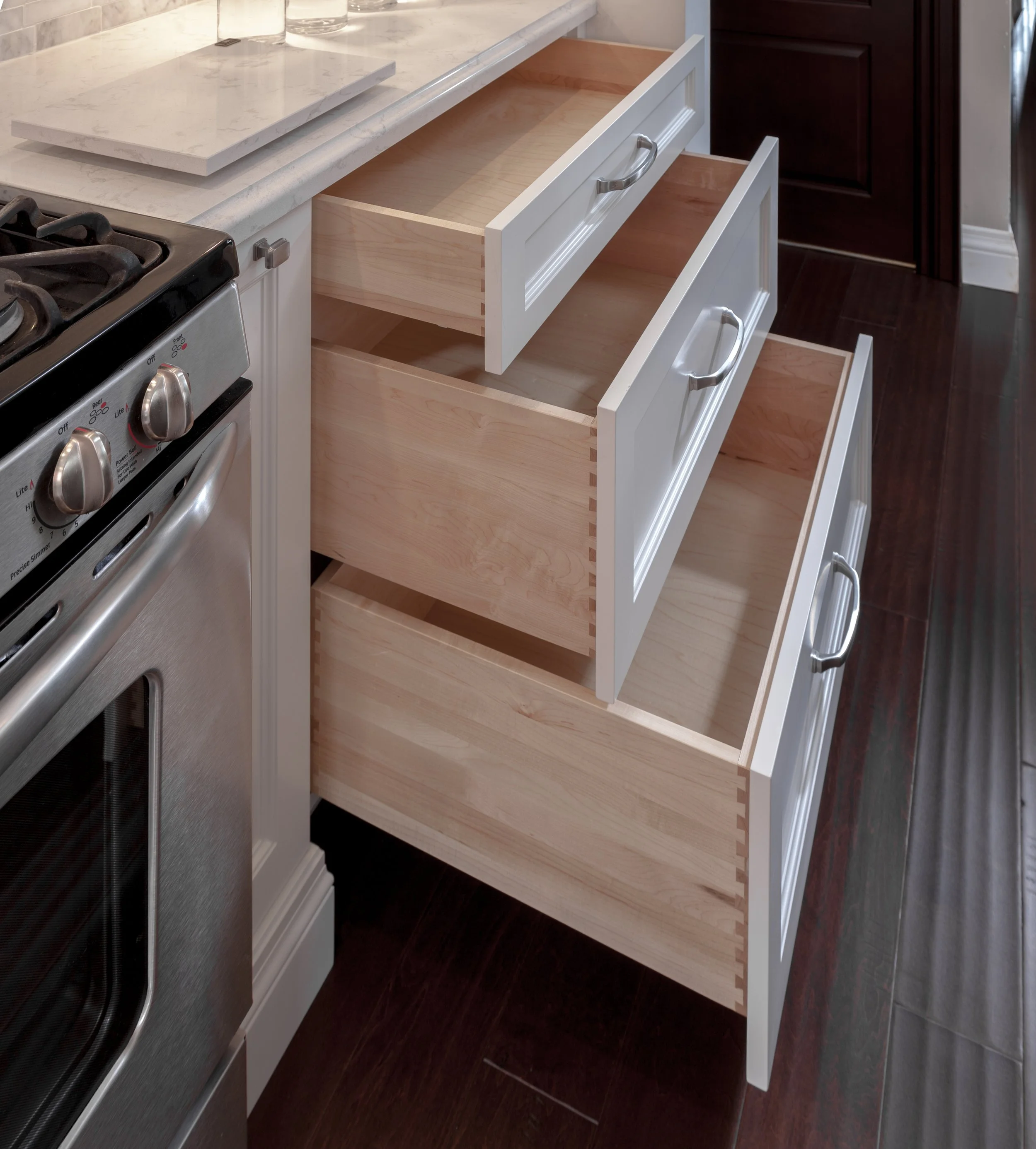 Empty kitchen cabinet drawers with white fronts and silver handles, situated under a marble countertop next to a stove, with dark wood flooring and cabinets nearby.