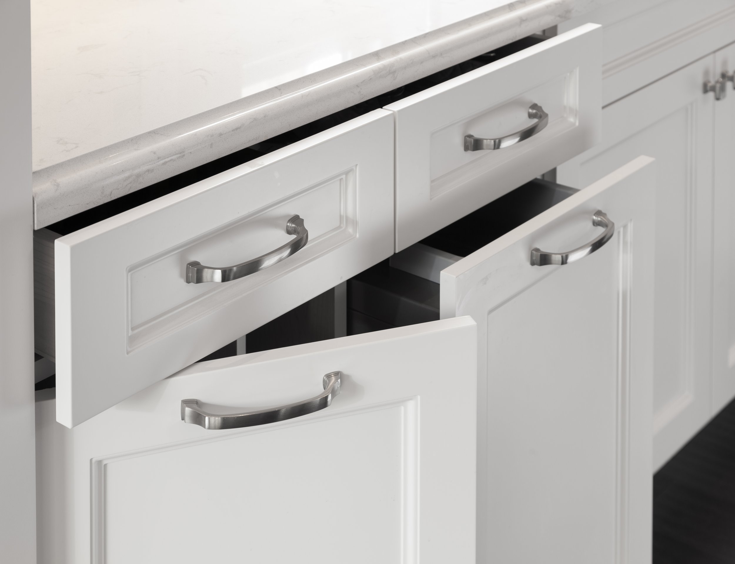 White kitchen cabinet drawers with silver handles, partially open, below a marble countertop.