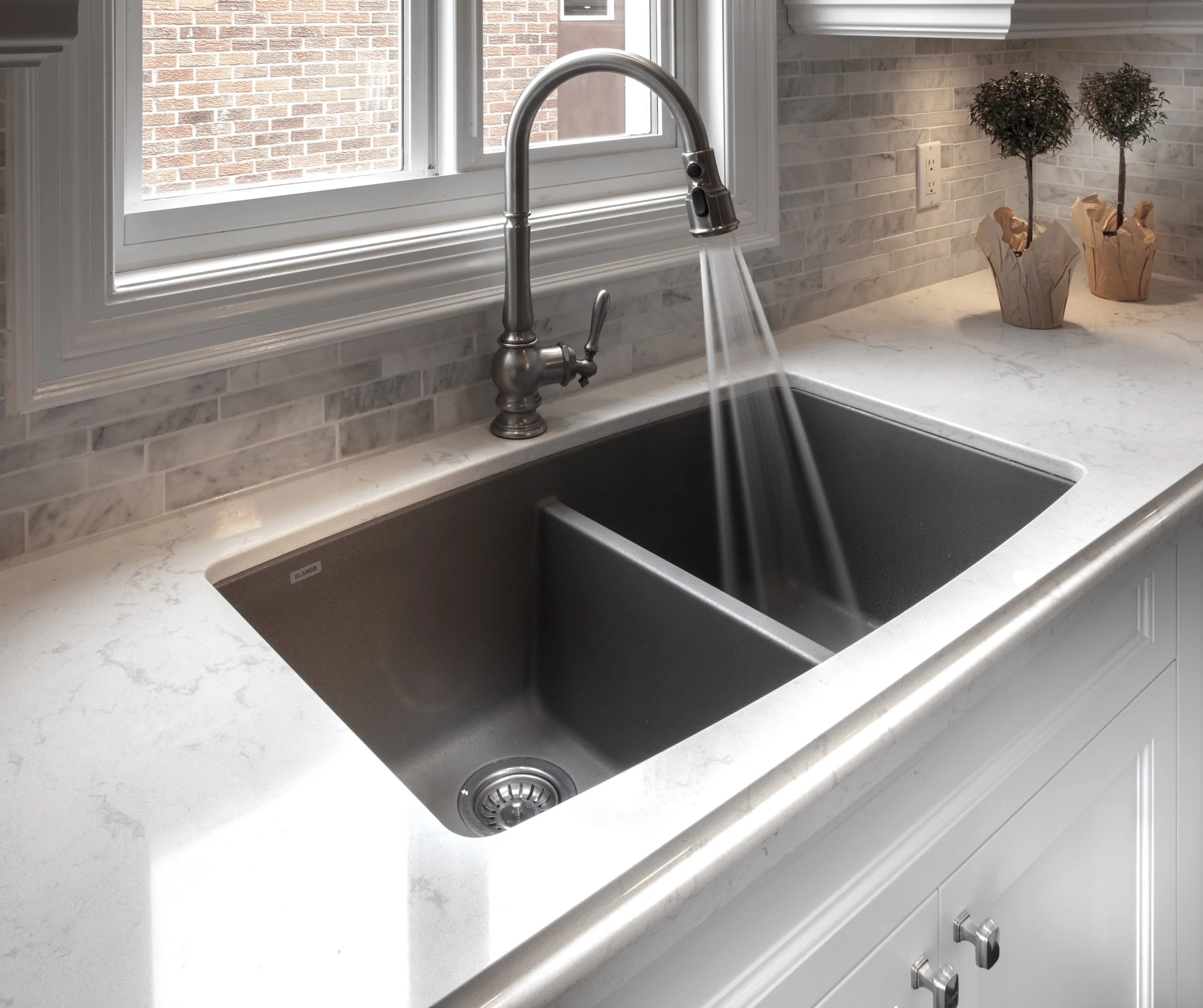 A kitchen sink with a stainless steel faucet and a white marble countertop near a window, with decorative plants in paper bags on the right side.