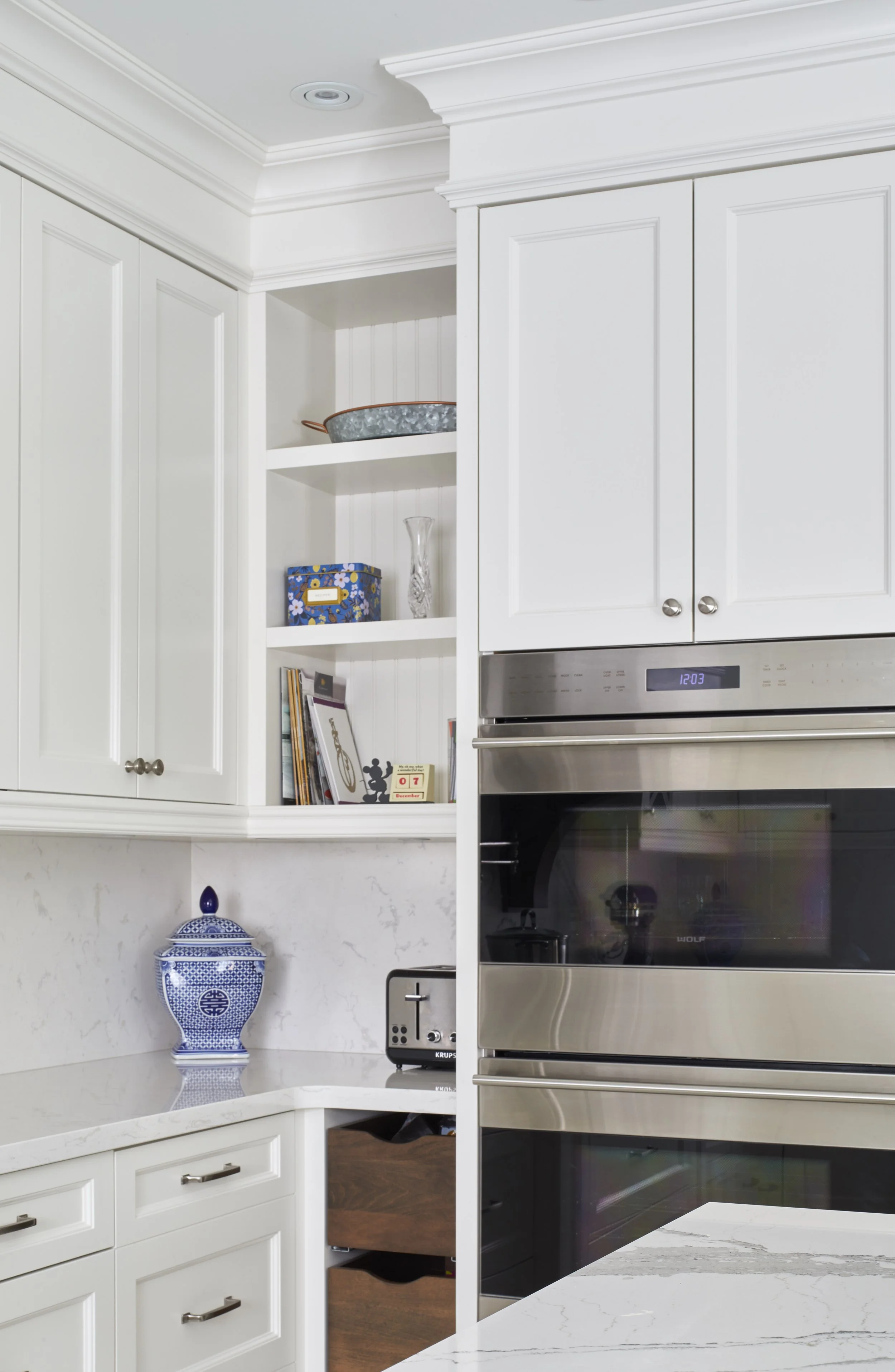 White kitchen with white cabinetry, stainless steel oven, blue and white decorative jar, open shelves with books and decorative items, marble countertop.