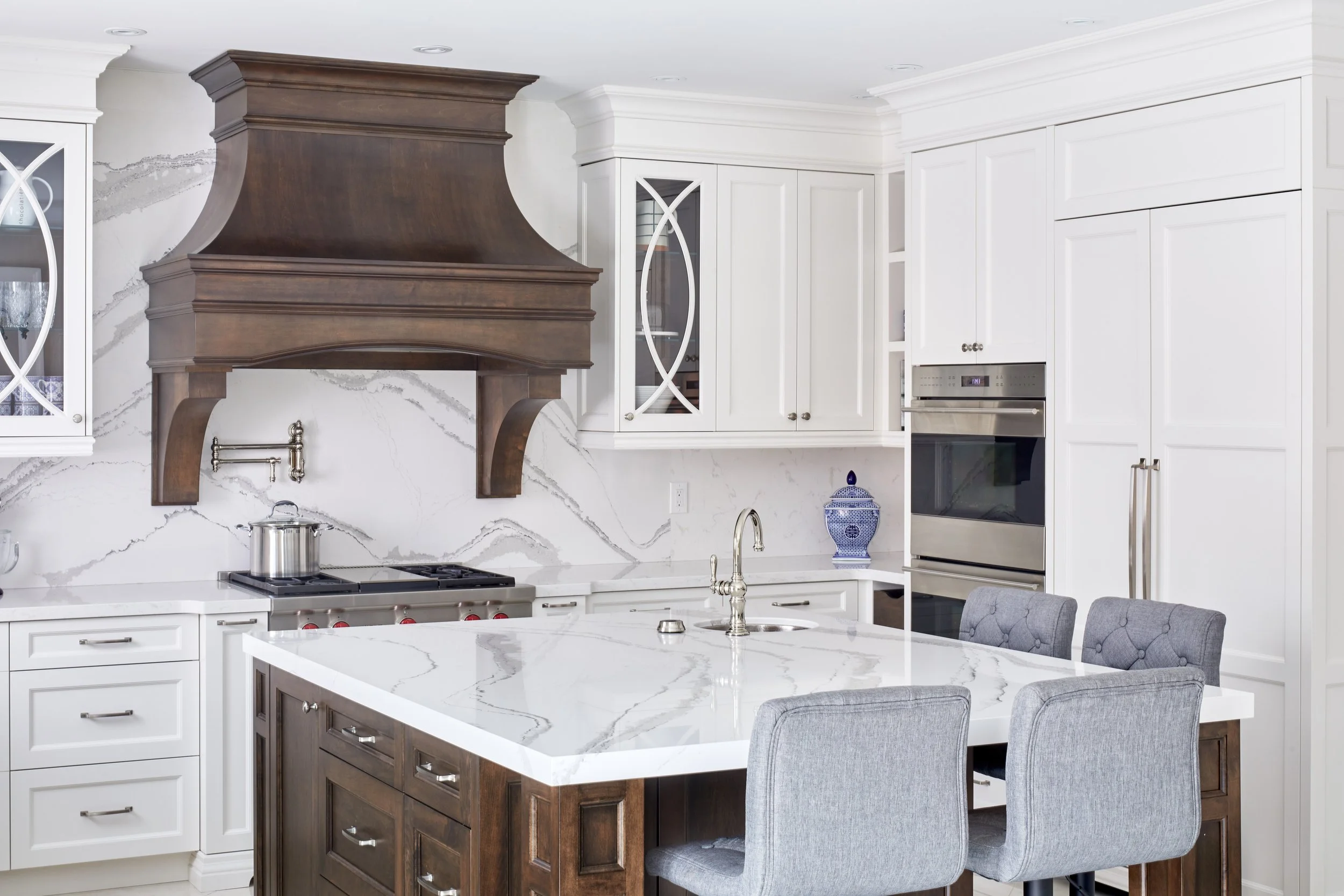 Modern kitchen with white cabinets, a marble island, and a wooden range hood above a cooktop. Gray upholstered chairs are placed around the island. There is a blue and white decorative jar on the counter and a built-in oven.