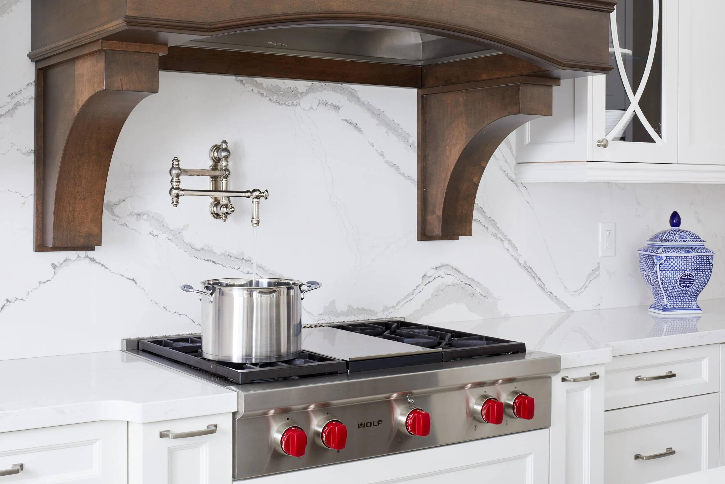 Kitchen stove and countertop with a pot on the burner, white cabinets, a decorative blue and white jar, and a marble backsplash.