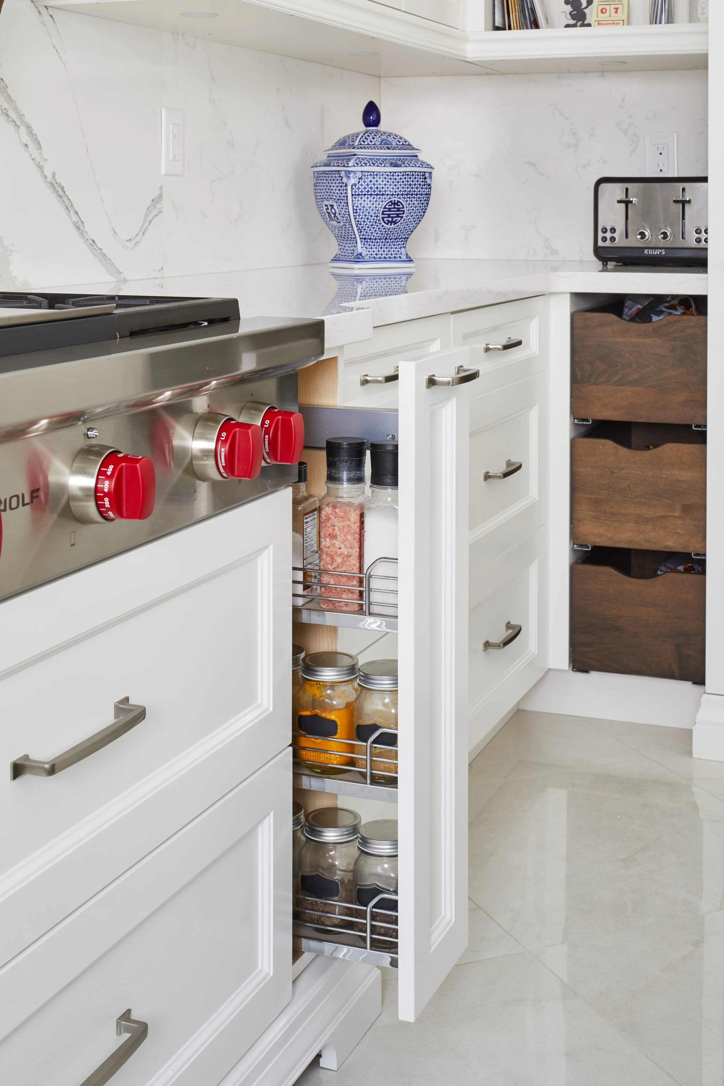 A kitchen with white cabinetry, a marble backsplash, a blue and white decorative vase on the counter, a stove with red control knobs, and pull-out spice jars visible inside a cabinet.
