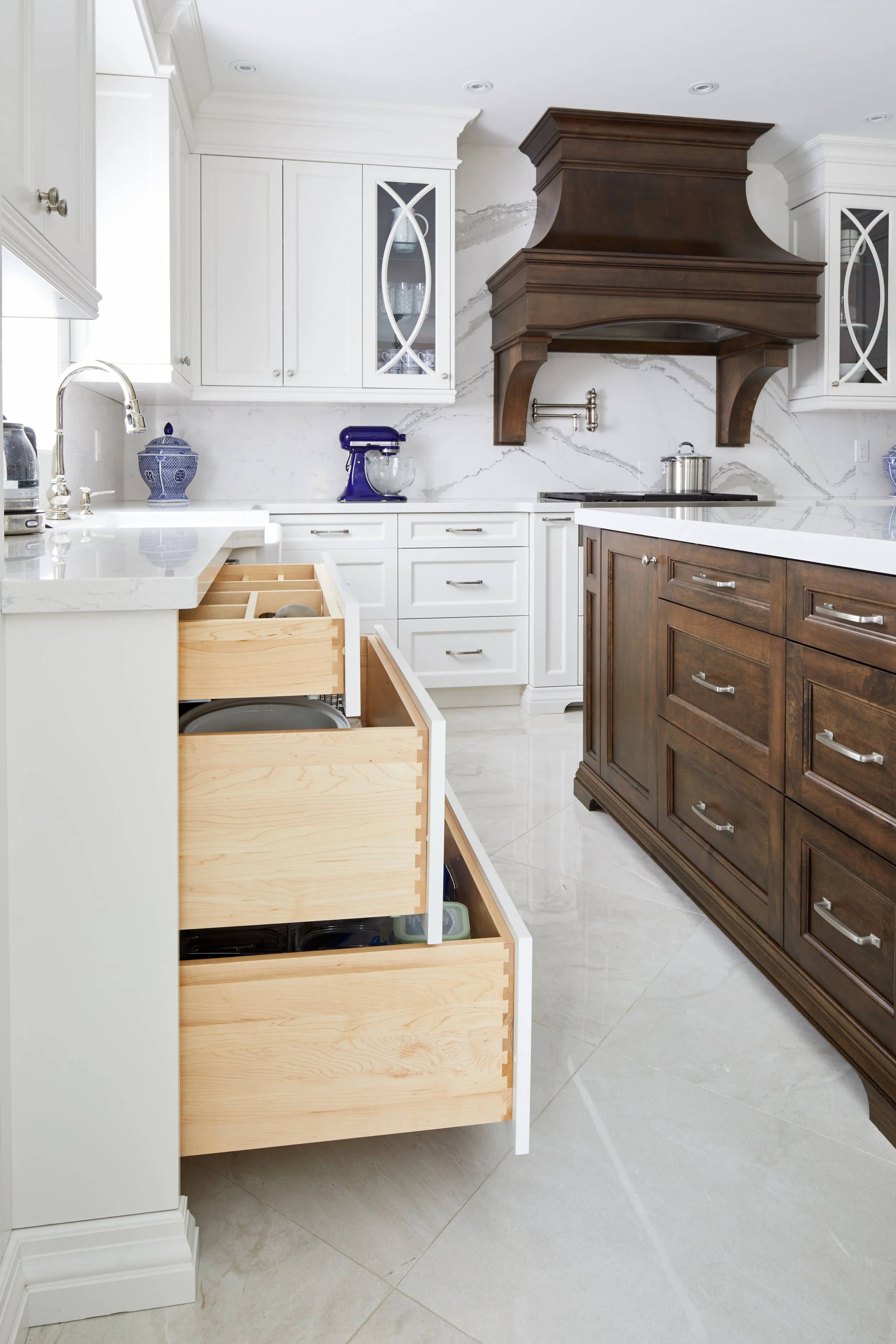Modern kitchen with white cabinets, a marble backsplash, dark wooden range hood, and partially open drawers on a light-colored tiled floor.