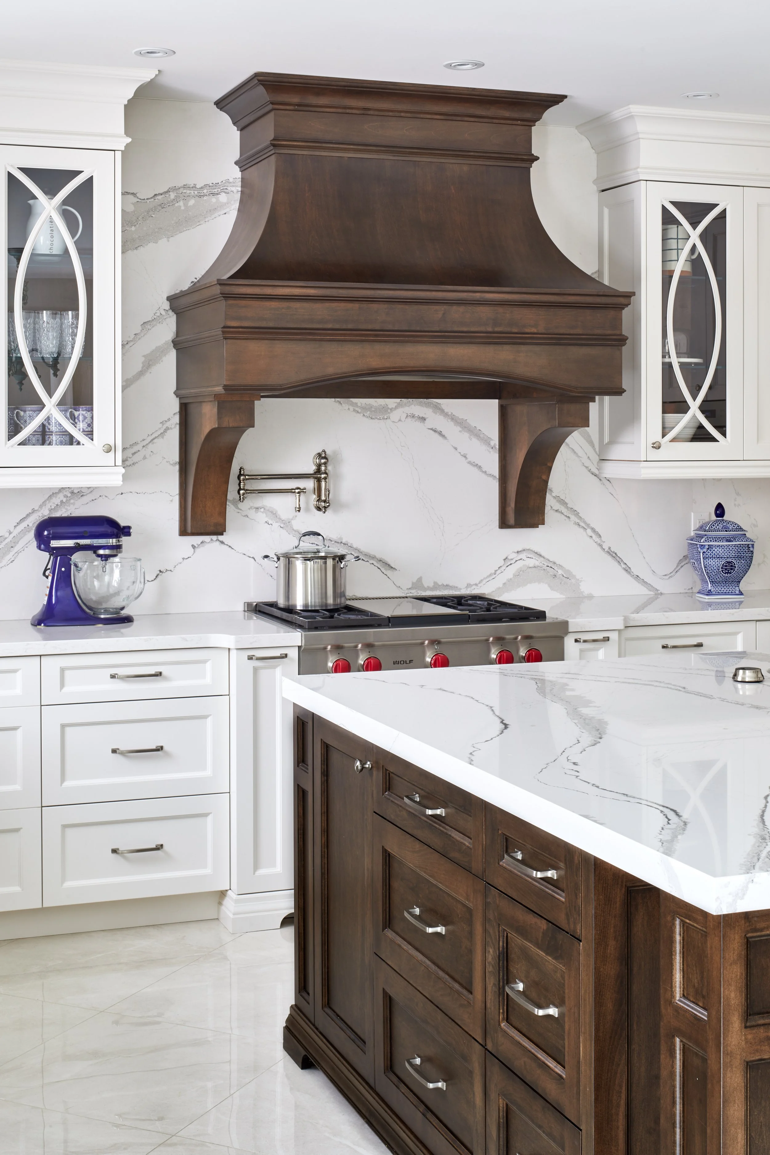 A modern kitchen with a marble island countertop, white and dark wooden cabinets, a stainless steel stove, and a dark wooden range hood. There are white cabinets with glass doors on either side of the range hood, and decorative items inside.