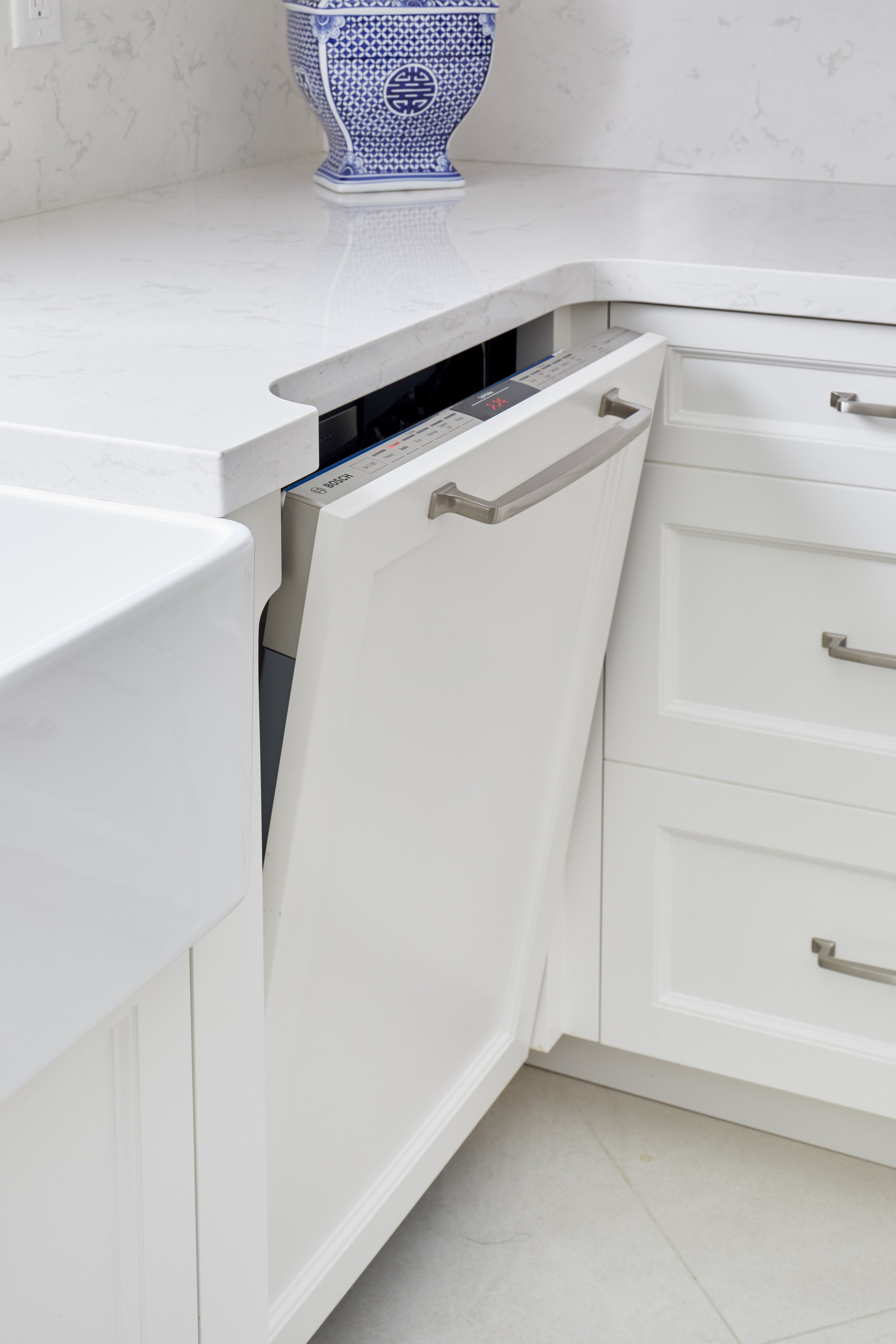 White kitchen cabinet with a built-in dishwasher, white countertop, and a blue and white ceramic vase on the countertop.