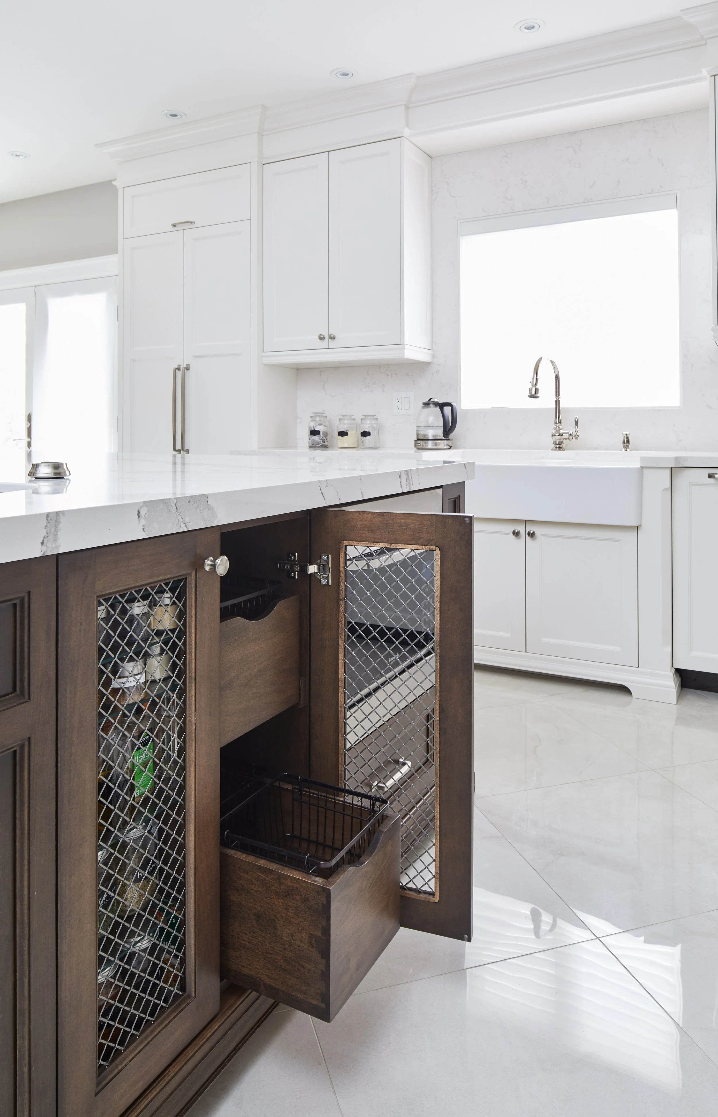 Kitchen with white cabinets, a white marble countertop, and a large window above the sink. Dark wooden cabinet with open drawers in the foreground. Items include jars, a kettle, and various kitchen appliances.