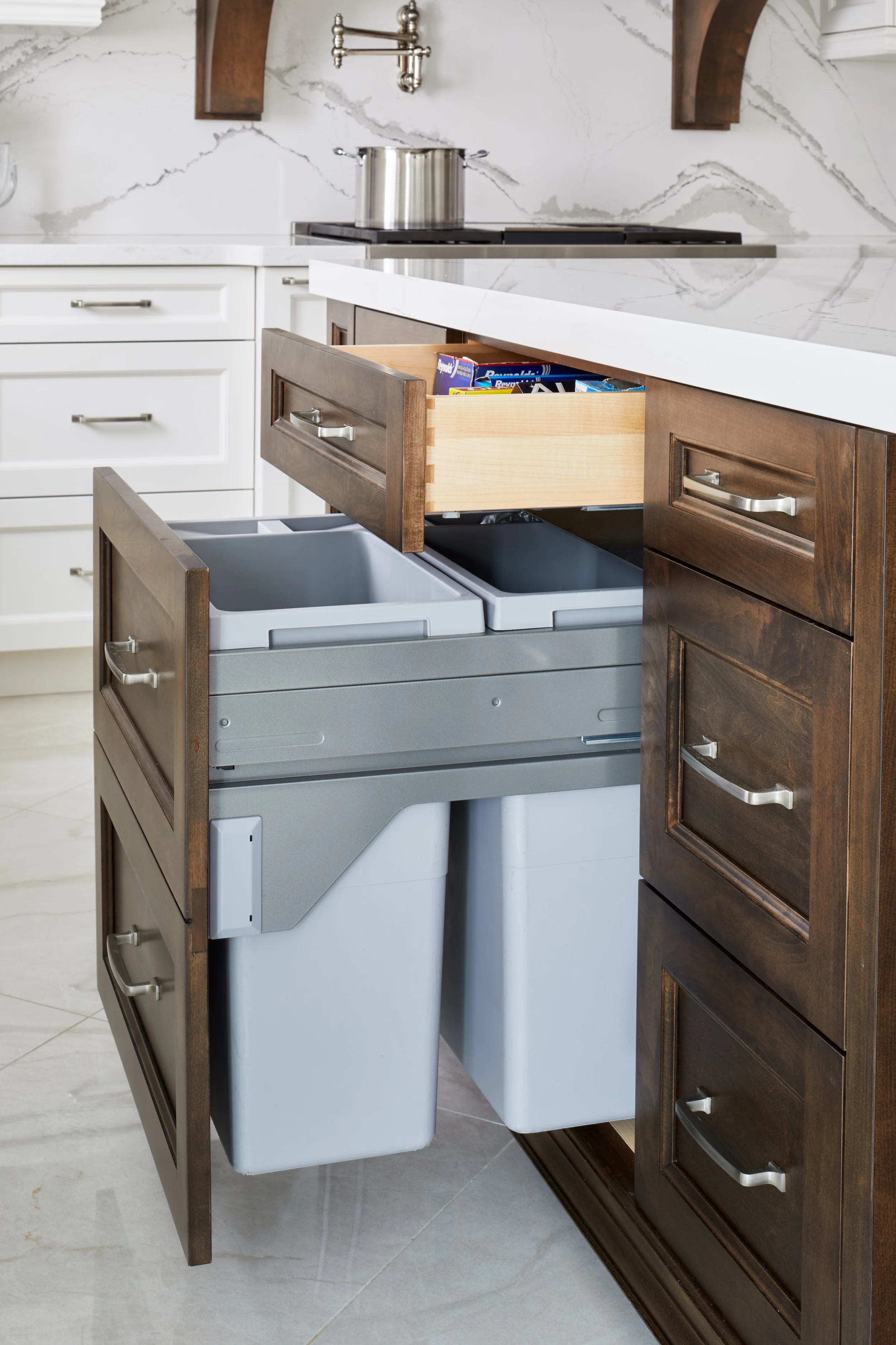 Kitchen with white cabinets, marble backsplash, and a wooden drawer pulled out of a dark wood cabinet, revealing organized recycling and trash bins.
