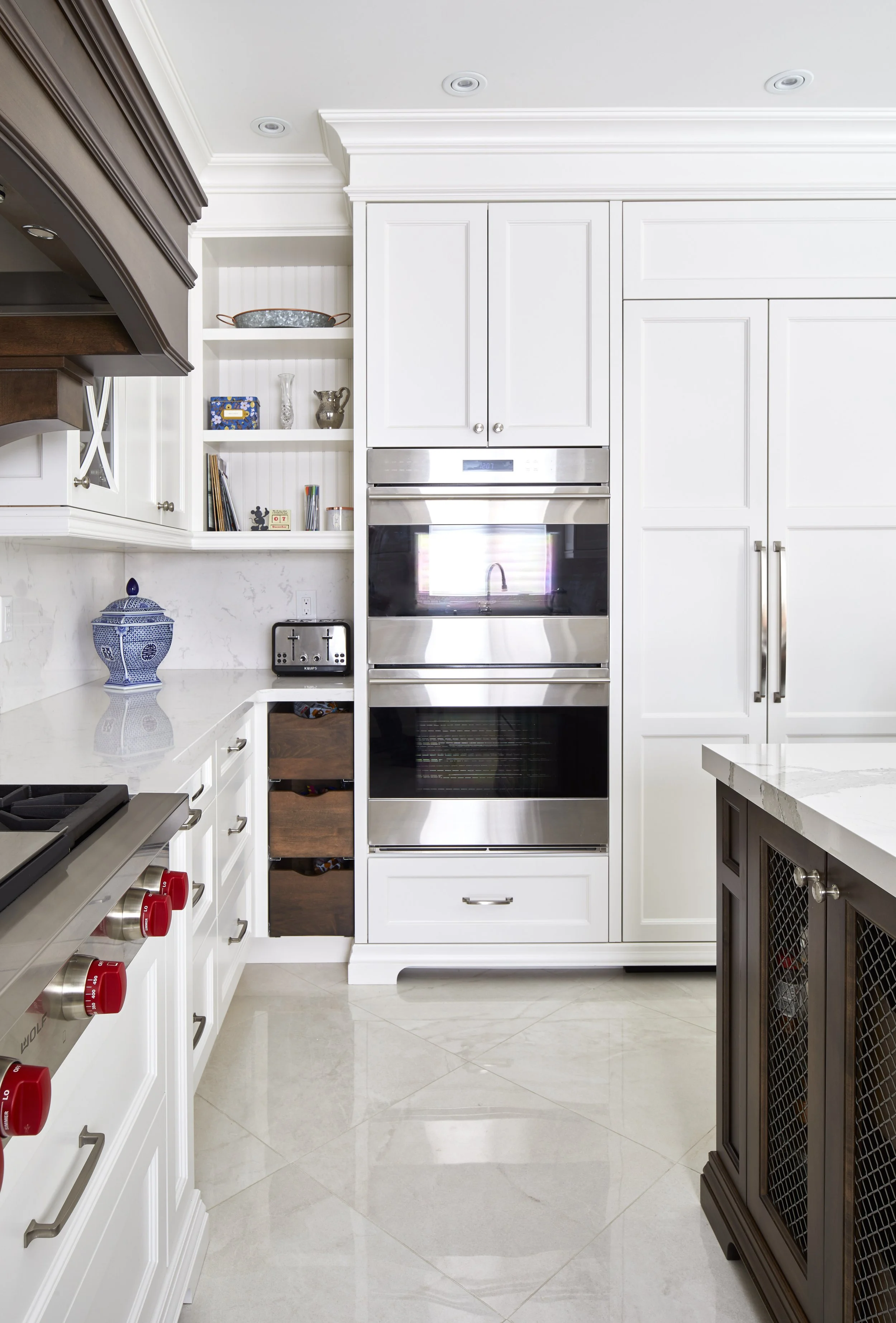 Modern kitchen with white cabinetry, stainless steel double oven, open shelves with decorative items, and a dark brown side cabinet, beige tiled floor.