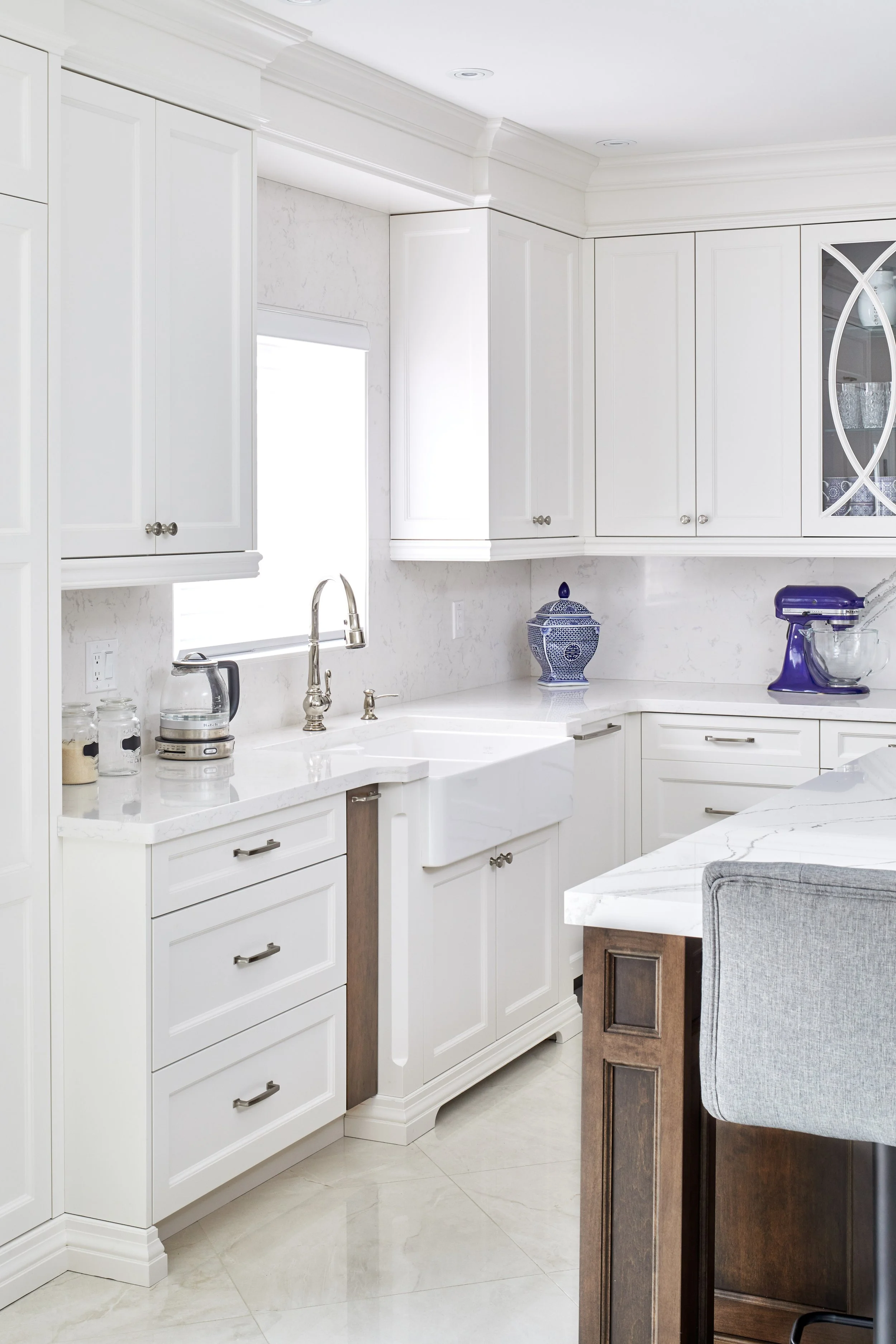 White kitchen with cabinetry, marble countertops, a stainless steel sink with a brush, decorative jars, and a blue and white ceramic container. A small section of a gray upholstered chair and a dark wooden table are visible.