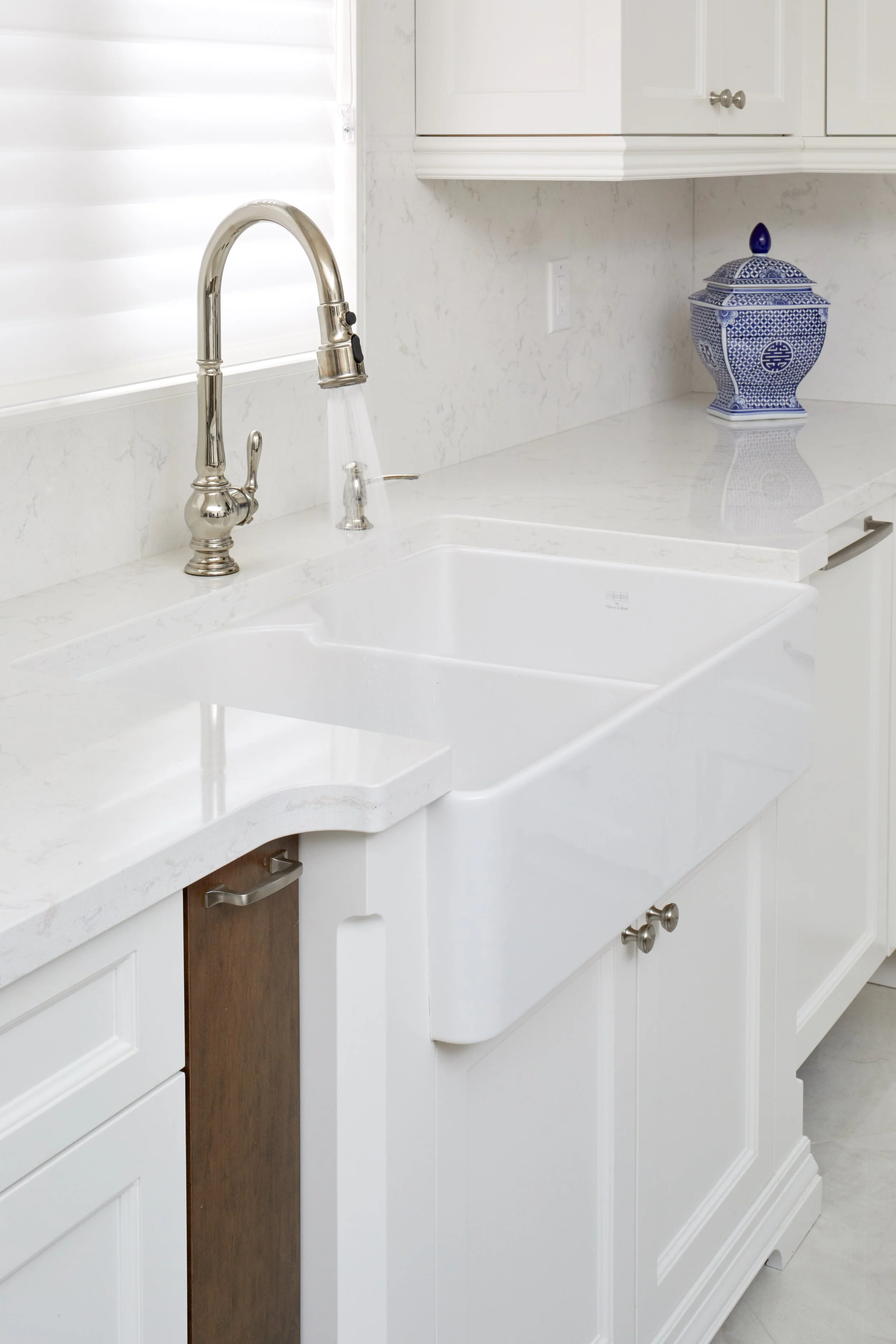 White kitchen counter with a silver faucet, blue and white decorative jar, white cabinets, and a window with white blinds.