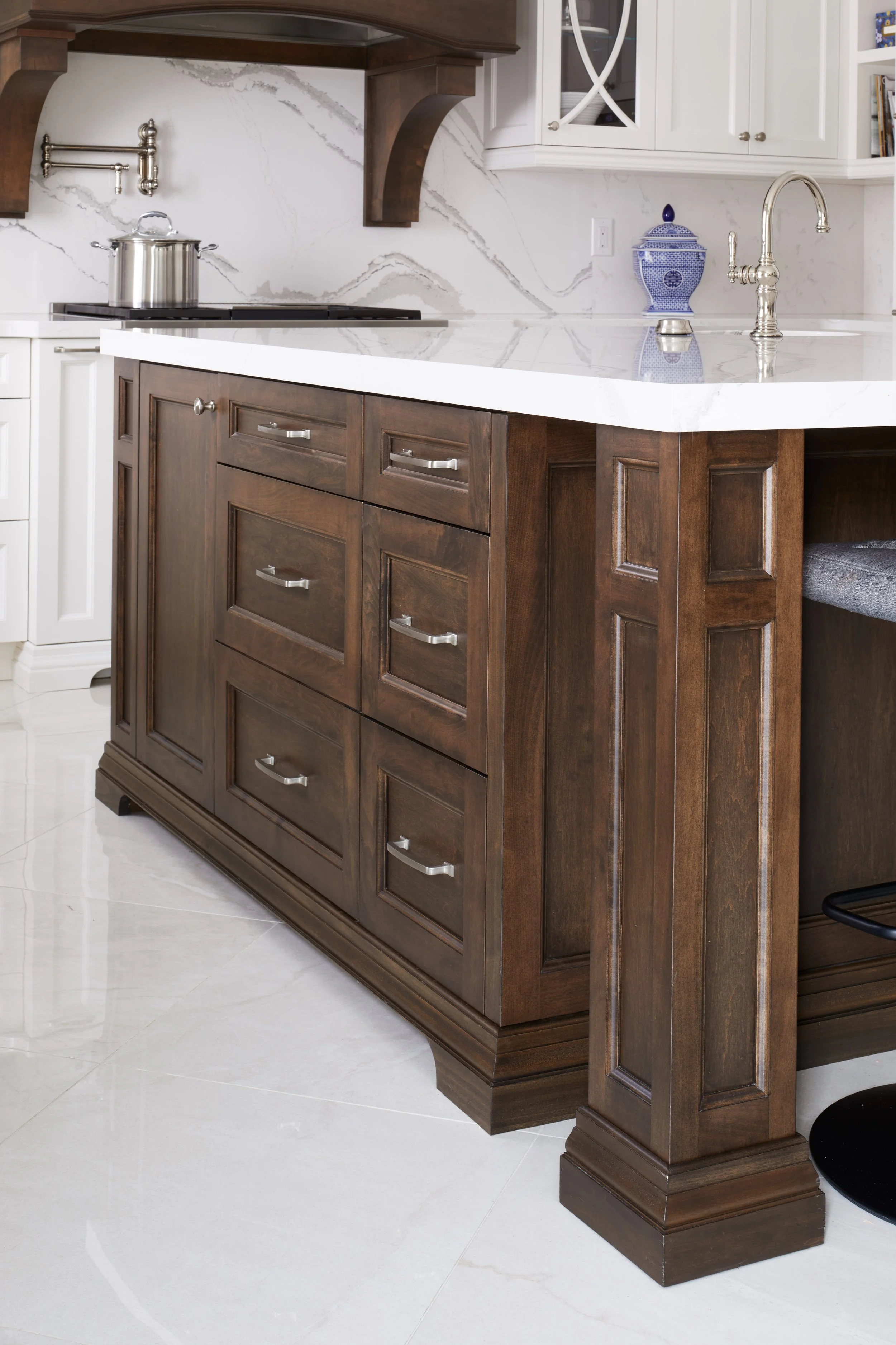 A kitchen island with dark wooden cabinets, a white marble countertop, and a shiny brass faucet. A blue and white porcelain jar is on the counter. The background shows white cabinets and a marble backsplash.