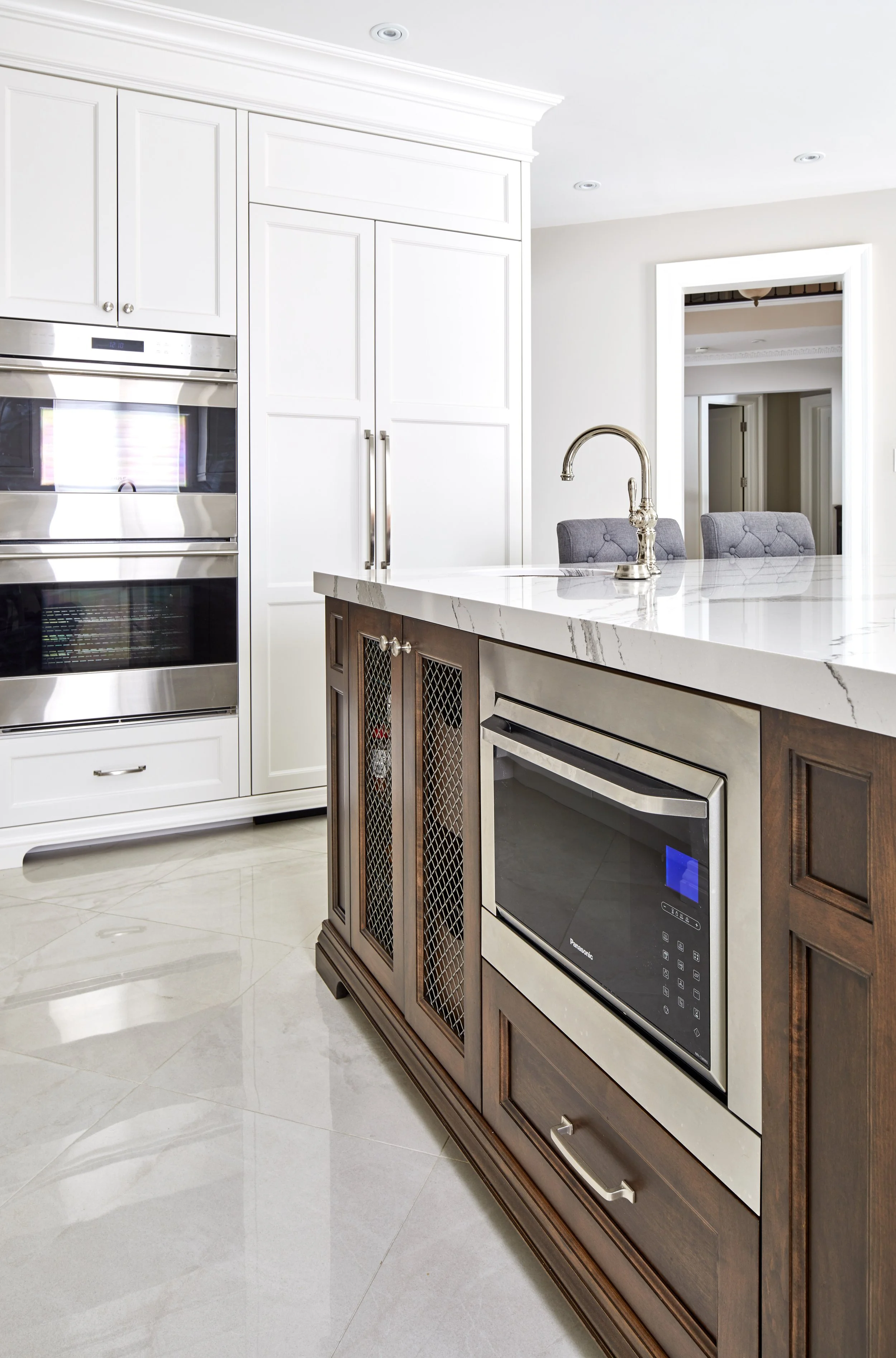 Modern kitchen with white cabinets, marble countertop, built-in microwave, and stainless steel appliances, including a double oven and a wine fridge. There's a marble island with a chrome faucet and cushioned chairs visible in the background.