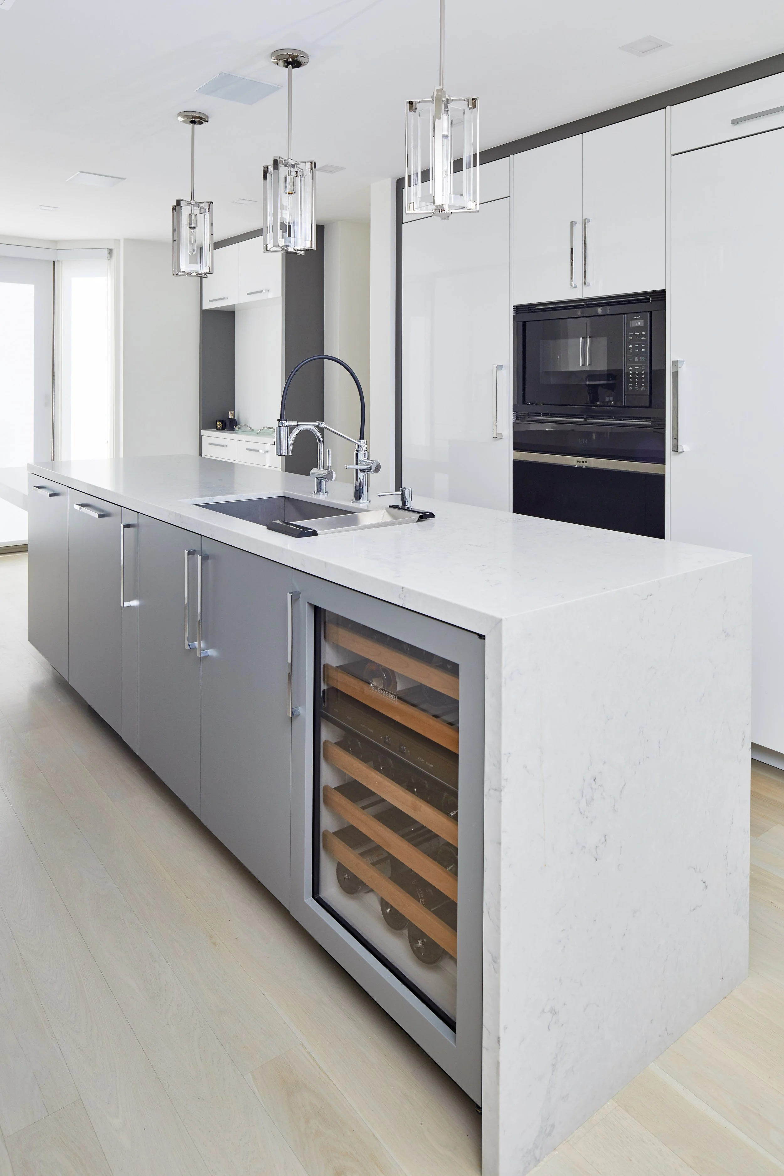 Modern kitchen with white and gray cabinetry, island with sink, black microwave oven, wine cooler with glass door, and three pendant lights with glass and metal fixtures hanging from the ceiling.