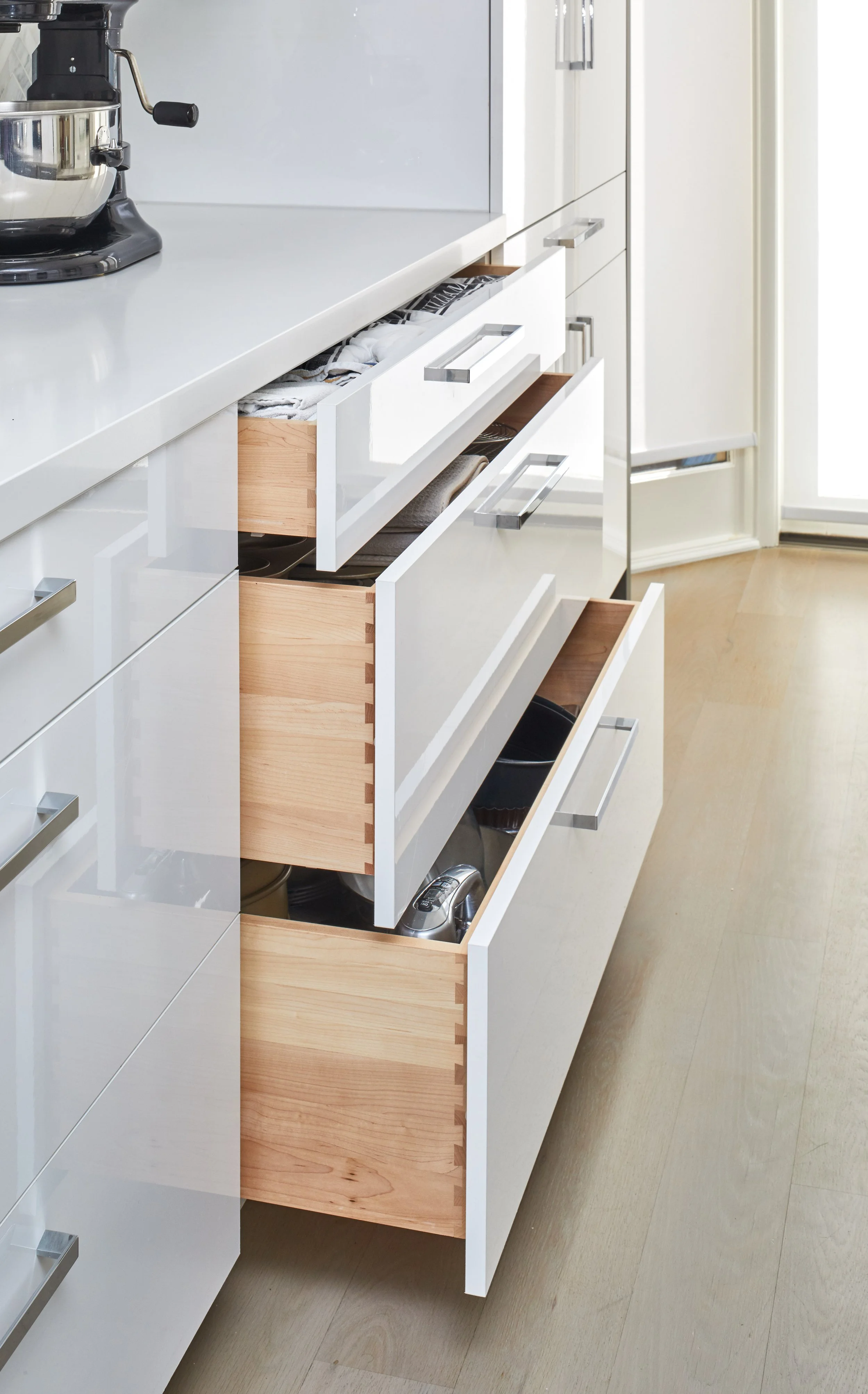 Open kitchen drawers, revealing stacked cookware and linens inside. The cabinet fronts are white with modern silver handles, and the floor is light wood.