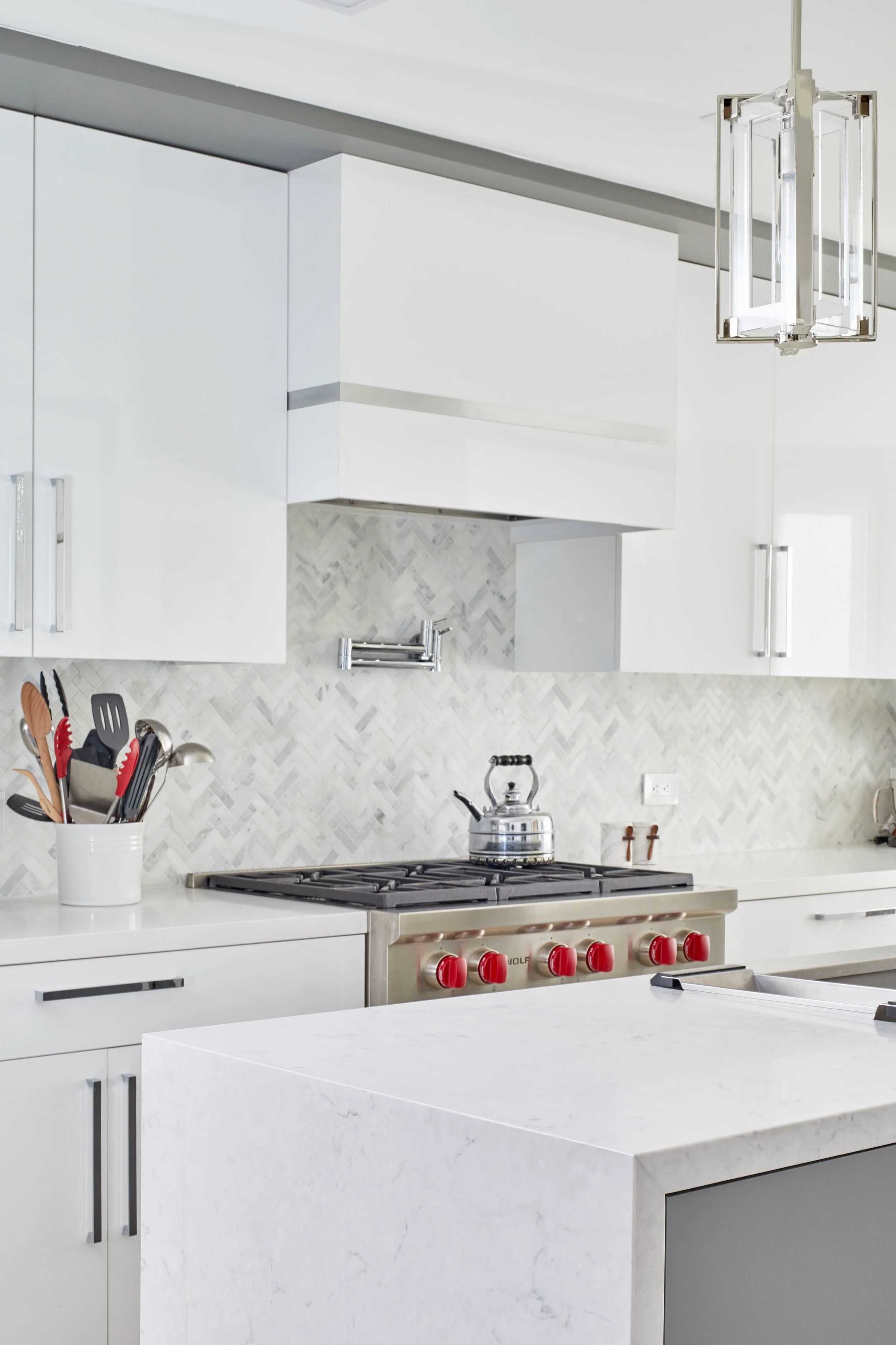 Modern white kitchen with a gas stove, a kettle on the stove, white cabinets, and kitchen utensils in a container. A pendant light hangs from the ceiling, and a marble-tile backsplash is behind the stove.