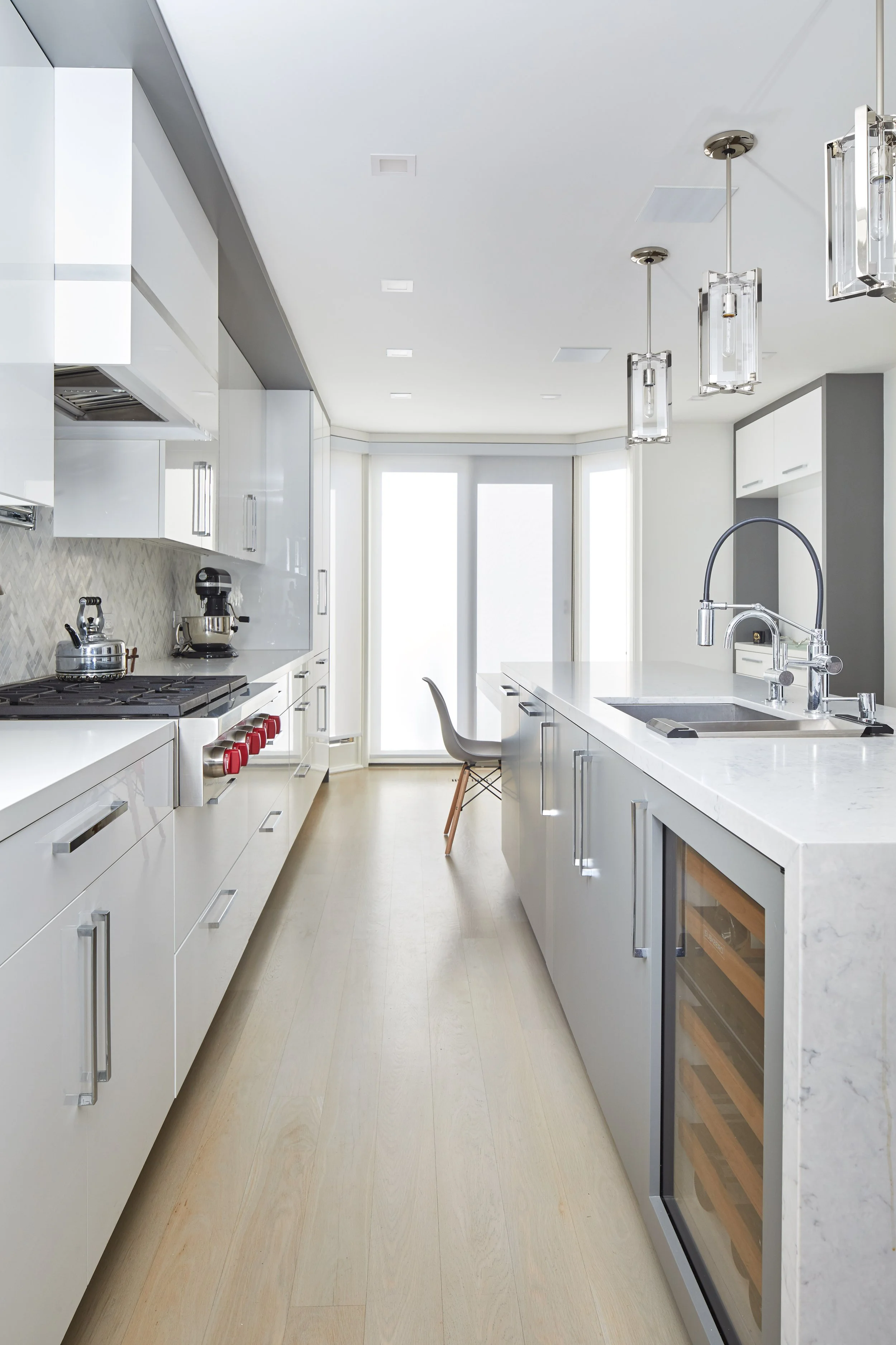 Modern white kitchen with island, stainless steel appliances, coffee maker, kettle, pendant lighting, and a wine cooler, bathed in natural light from large windows.