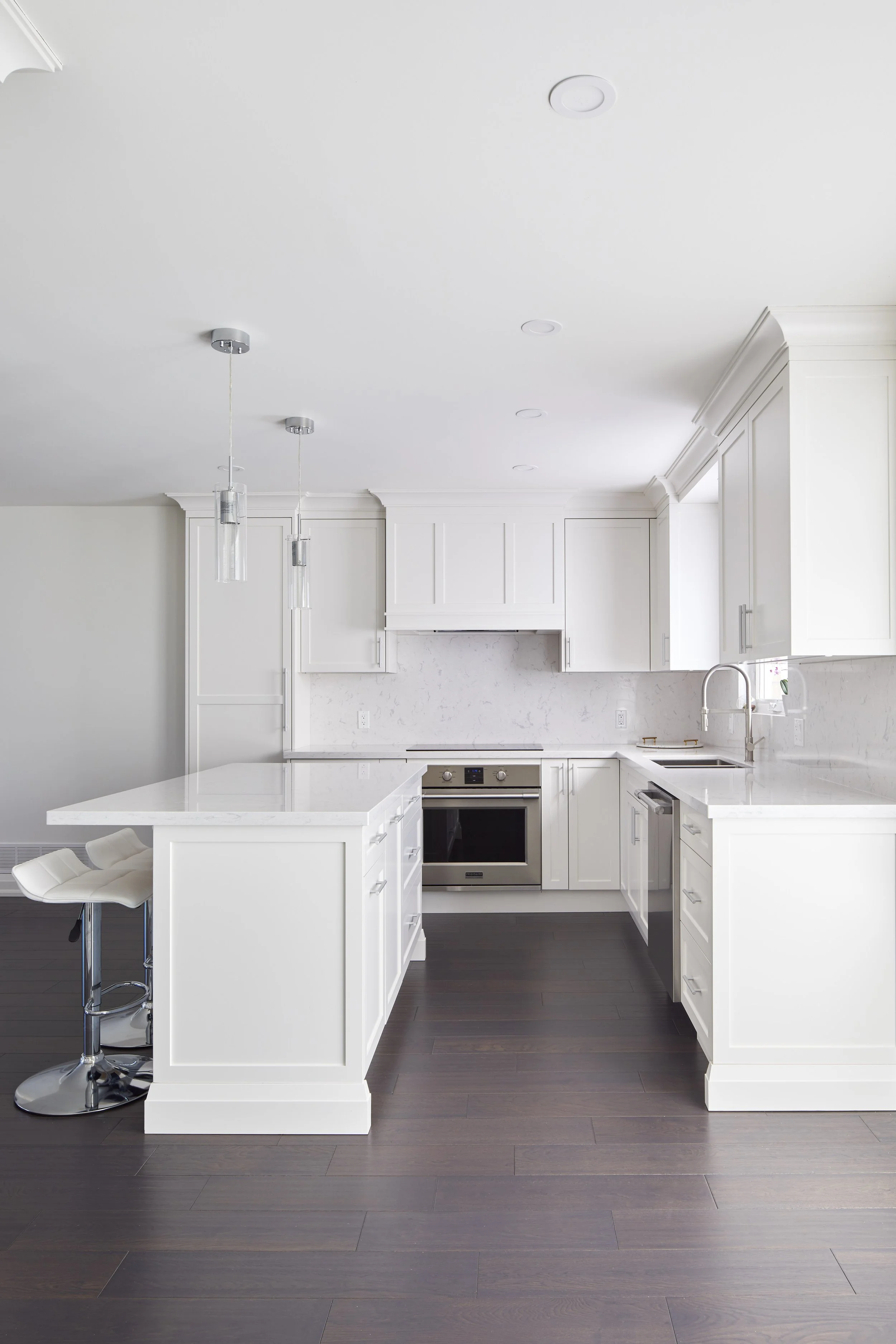 Modern white kitchen with dark hardwood flooring, white cabinets, stainless steel appliances, a kitchen island with a white countertop, and a white high-back chair at the counter.