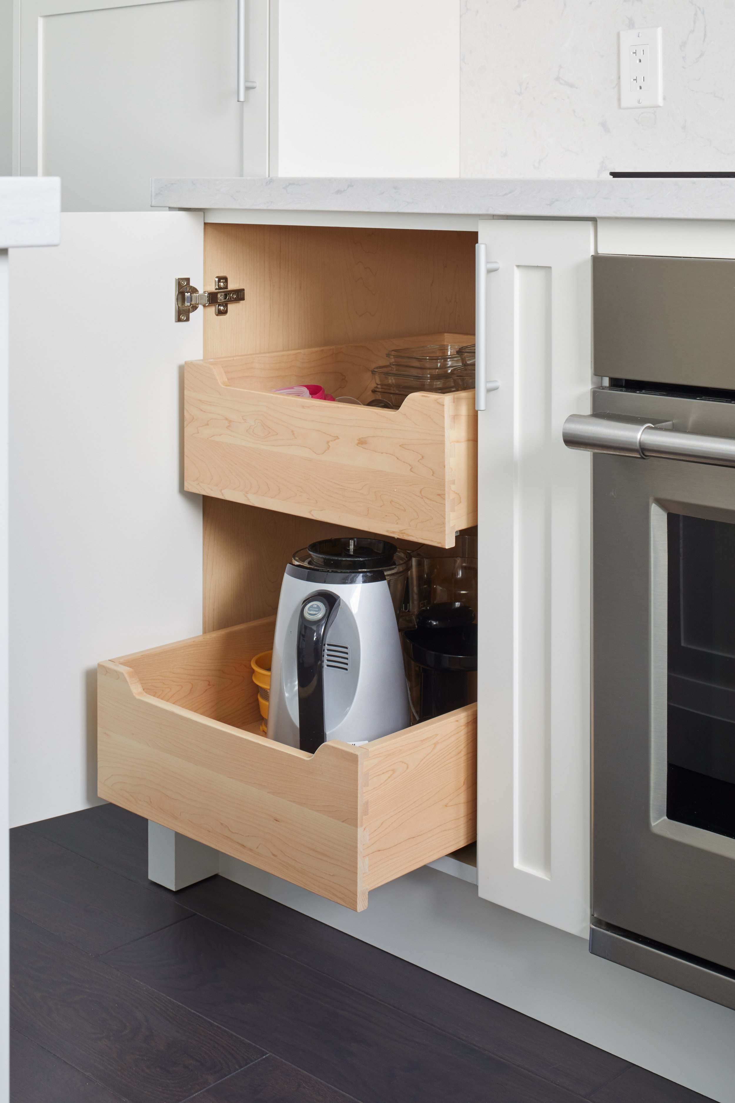 Open kitchen cabinet with three empty wooden drawers, containing a few small containers, kitchen utensils, and a blender on a dark hardwood floor.