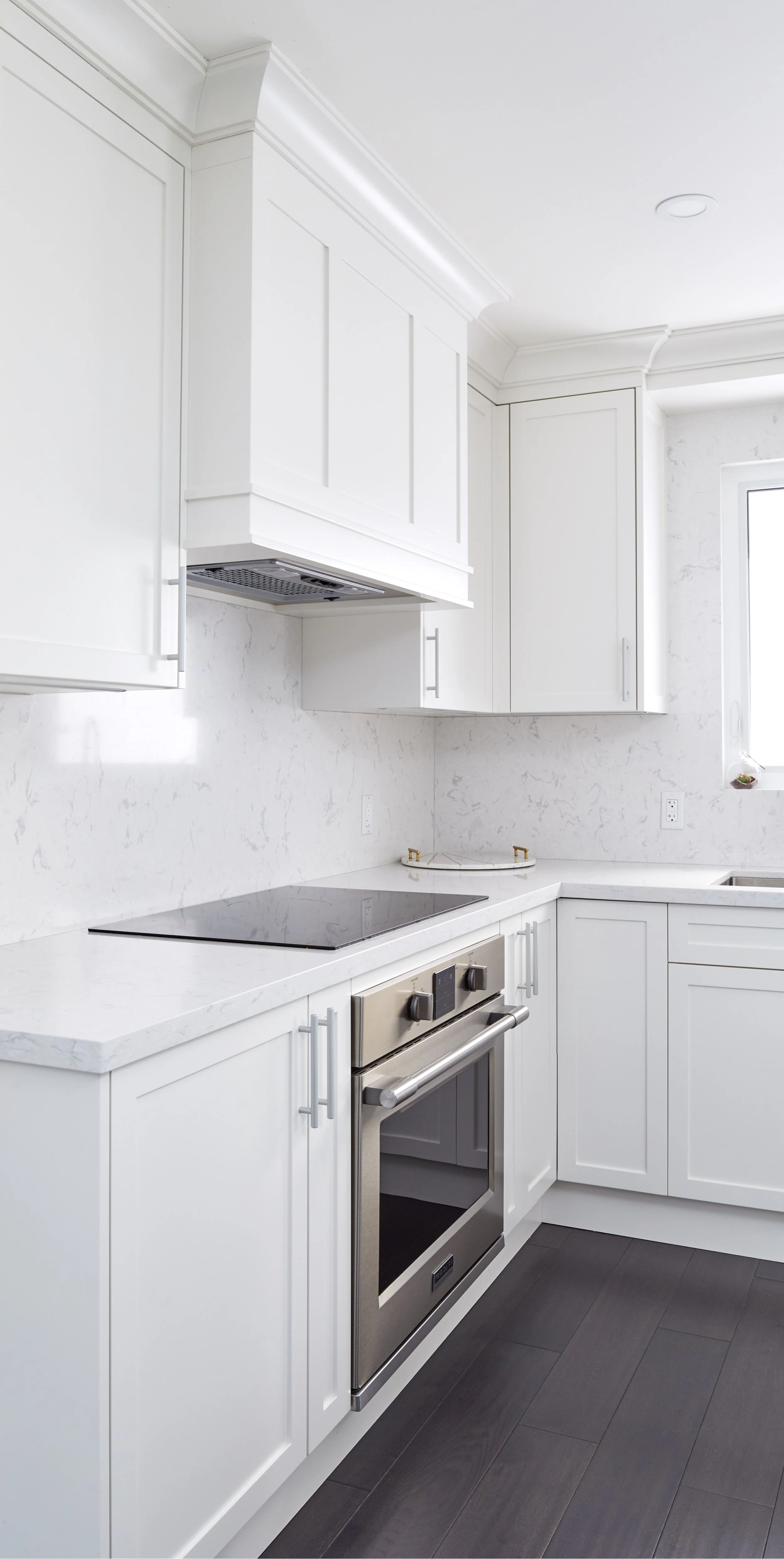A modern white kitchen with white cabinets, a marble backsplash, a black cooktop, stainless steel oven, and dark hardwood floors. Natural light coming through a window.