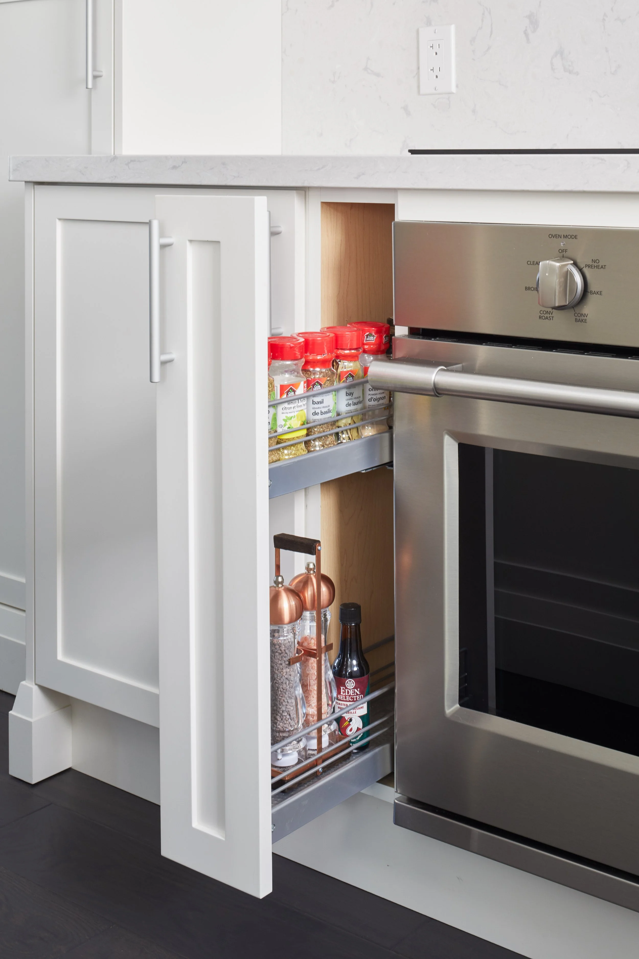 Open kitchen cabinet in white with spices and kitchen tools inside, next to a stainless steel oven.