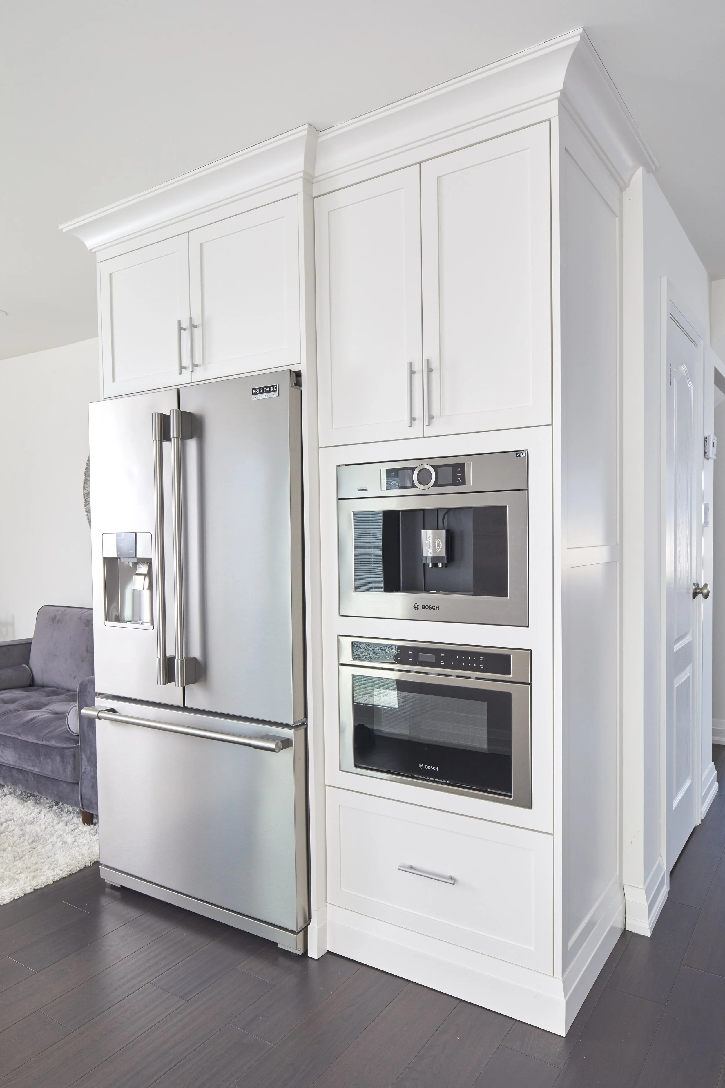 Kitchen with white cabinets and stainless steel refrigerator and oven, gray flooring, and a gray sofa in the background.
