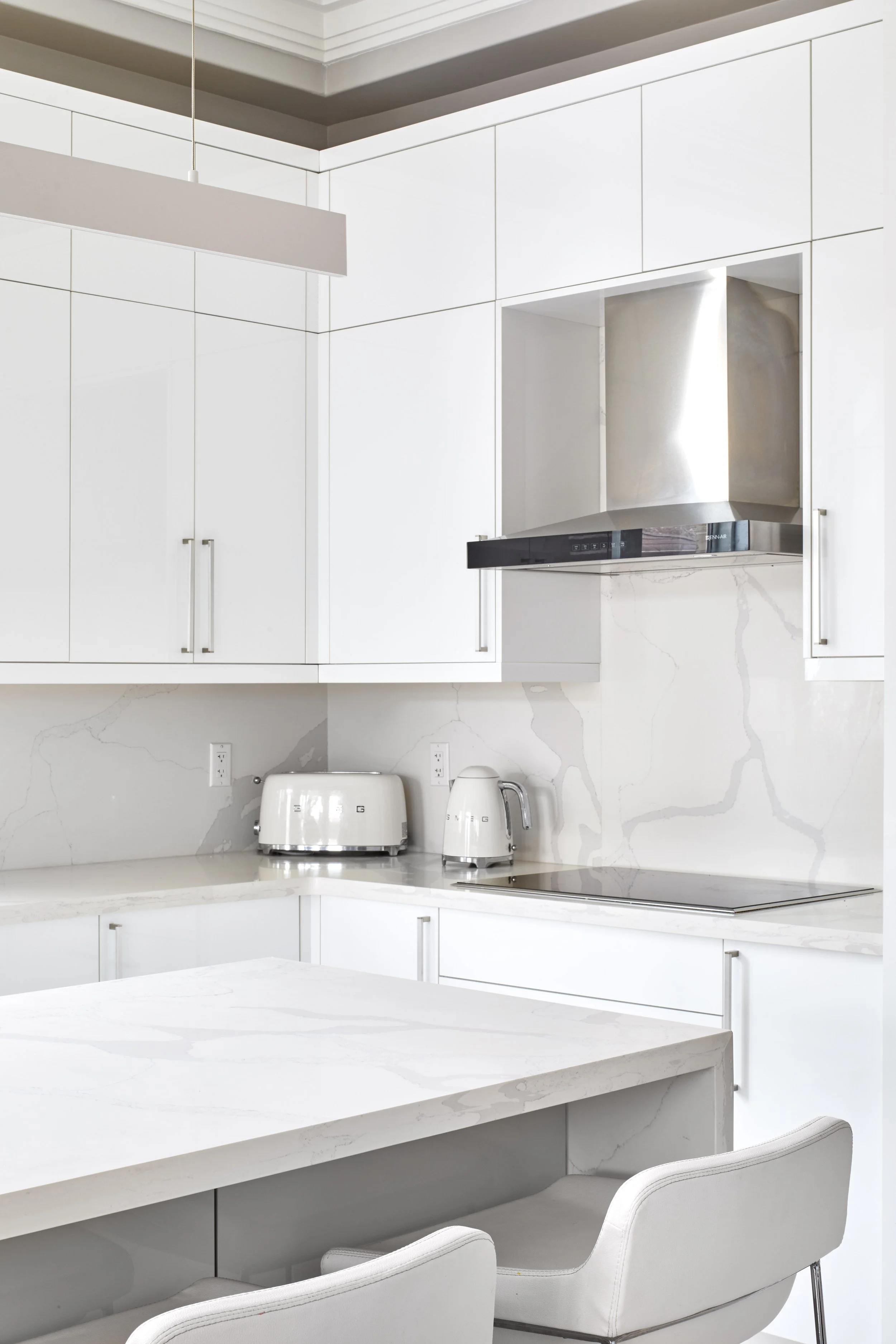 Modern kitchen with white cabinets, marble backsplash, and countertop; features a stainless steel range hood, toaster, electric kettle, and chairs.
